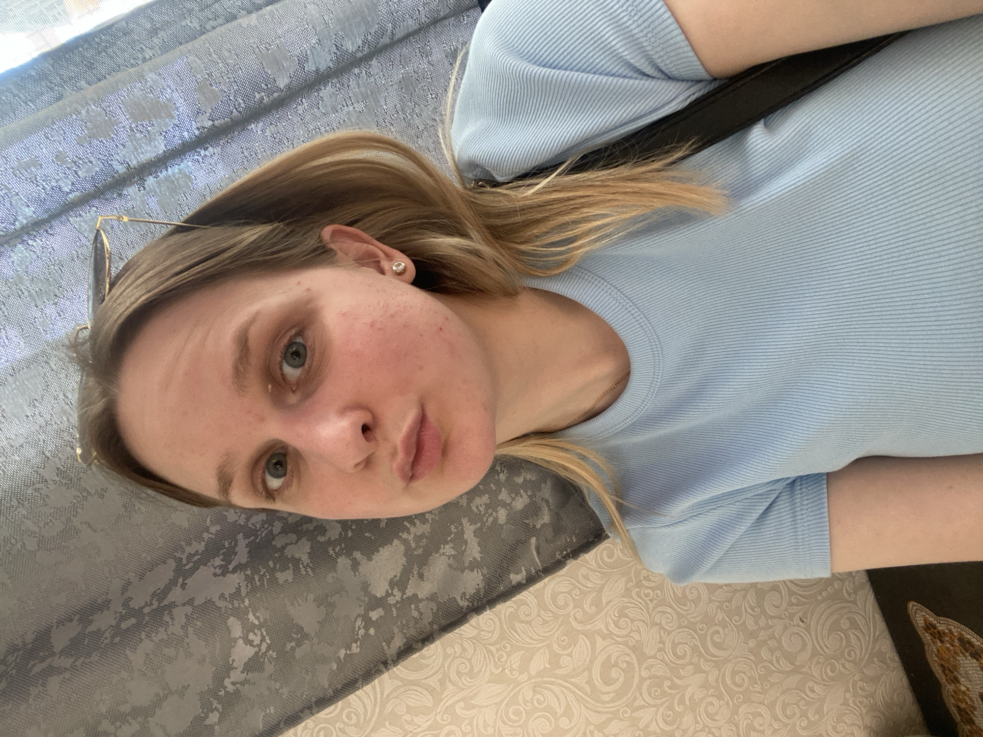 A young woman with blonde hair and blue eyes is shown in a close-up, self-taken photograph. She is wearing a light blue ribbed t-shirt and a silver stud earring in her left ear. Her hair is partially pulled back and she has sunglasses resting on her head. The background appears to be a home interior, with a patterned wallpaper on the left and a textured curtain on the right. The lighting suggests it is daytime. The image was taken in Chaikovsky, Russia.
