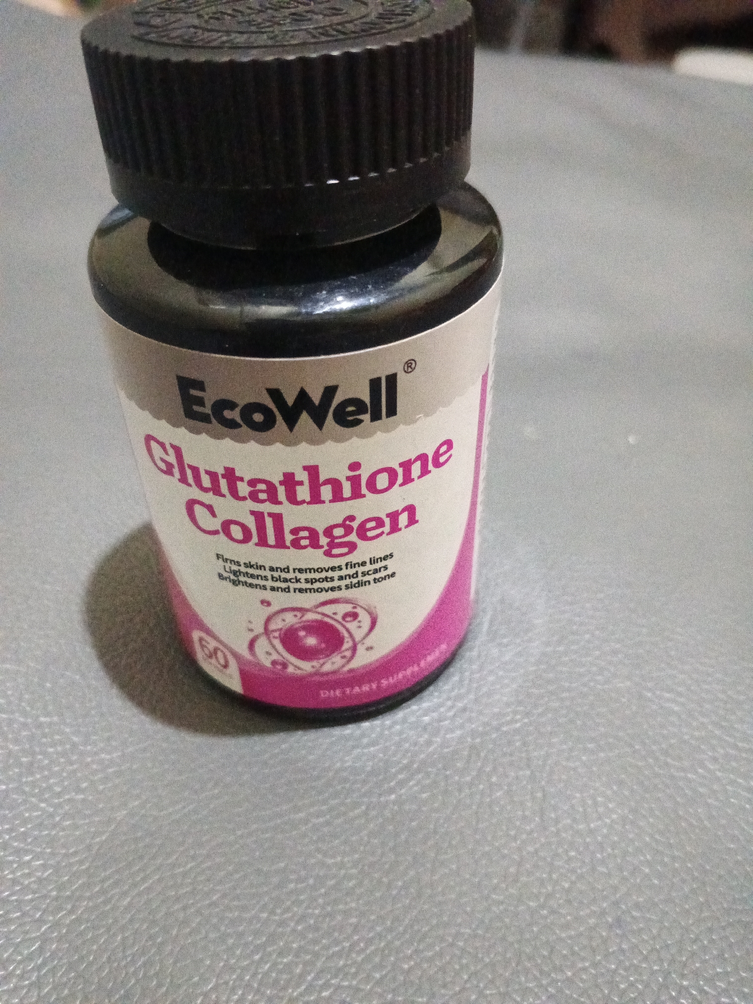 The image shows a close-up of a bottle of EcoWell Glutathione Collagen dietary supplement. The bottle has a black cap and a white label with pink text. The text on the label describes the product's benefits, such as firming skin, removing fine lines, lightening black spots and scars, and brightening skin tone. There is also a number "60" visible on the lower part of the label, likely indicating the number of capsules in the bottle. The bottle is placed on a textured grey surface. The background is blurred and out of focus, but appears to be an indoor setting. There are no people or other significant objects in the scene. The location in Penambangan, Indonesia is not discernible from the image itself.
