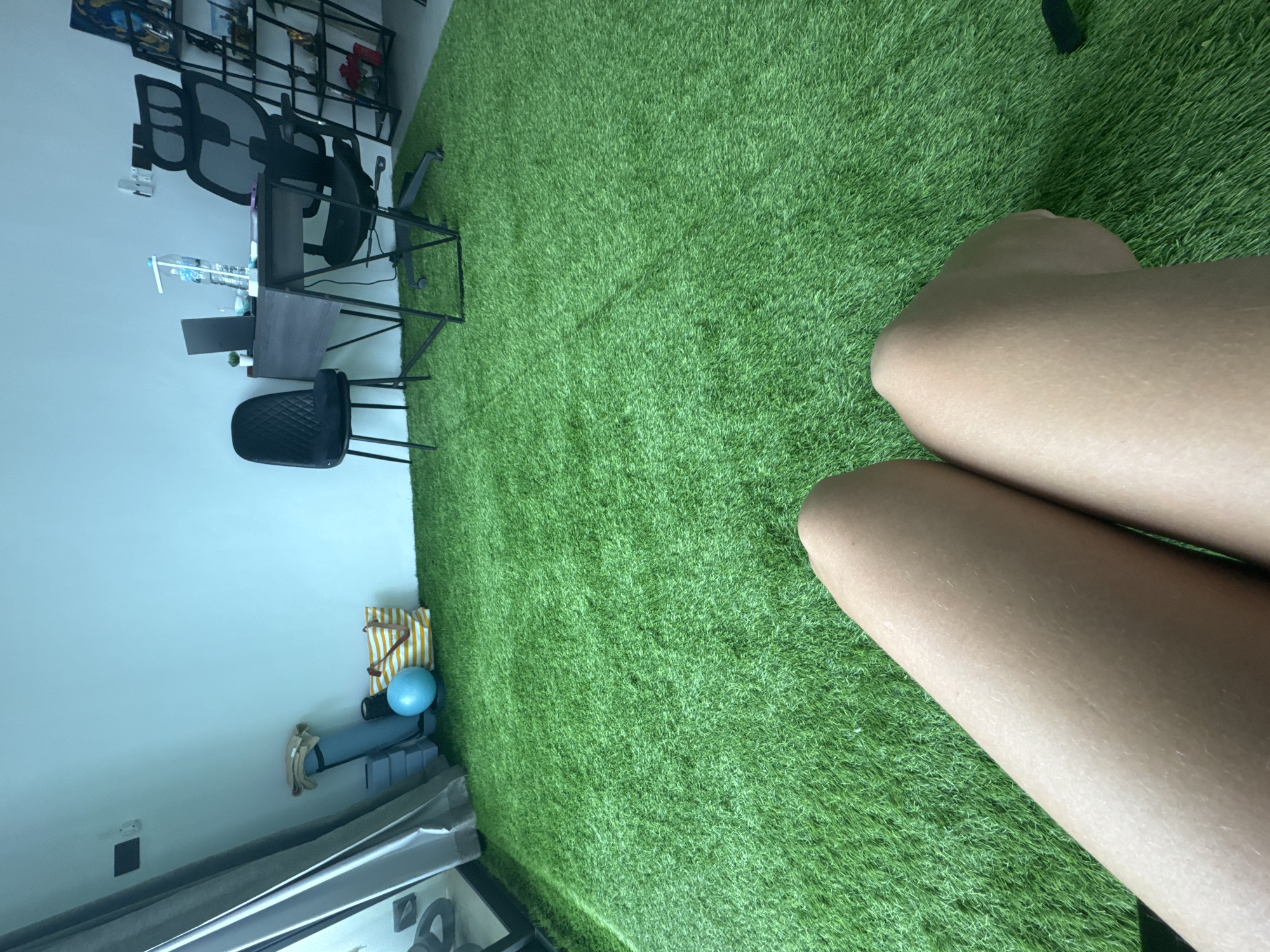 The image depicts an indoor space with artificial turf flooring, resembling a gym or recreational area, situated in a modern apartment or home in Koh Samui District, Thailand. In the foreground, the bare legs of a person are visible, positioned on the green turf. The background features a workspace with a dark desk and chairs. On the left side of the image, near a window with draped curtains, are exercise mats, a yoga mat, a blue ball, and a striped tote bag. The wall is painted a light blue. Further to the right, a shelf unit displays various items. The overall impression is one of a multi-functional living space within a tropical urban setting.