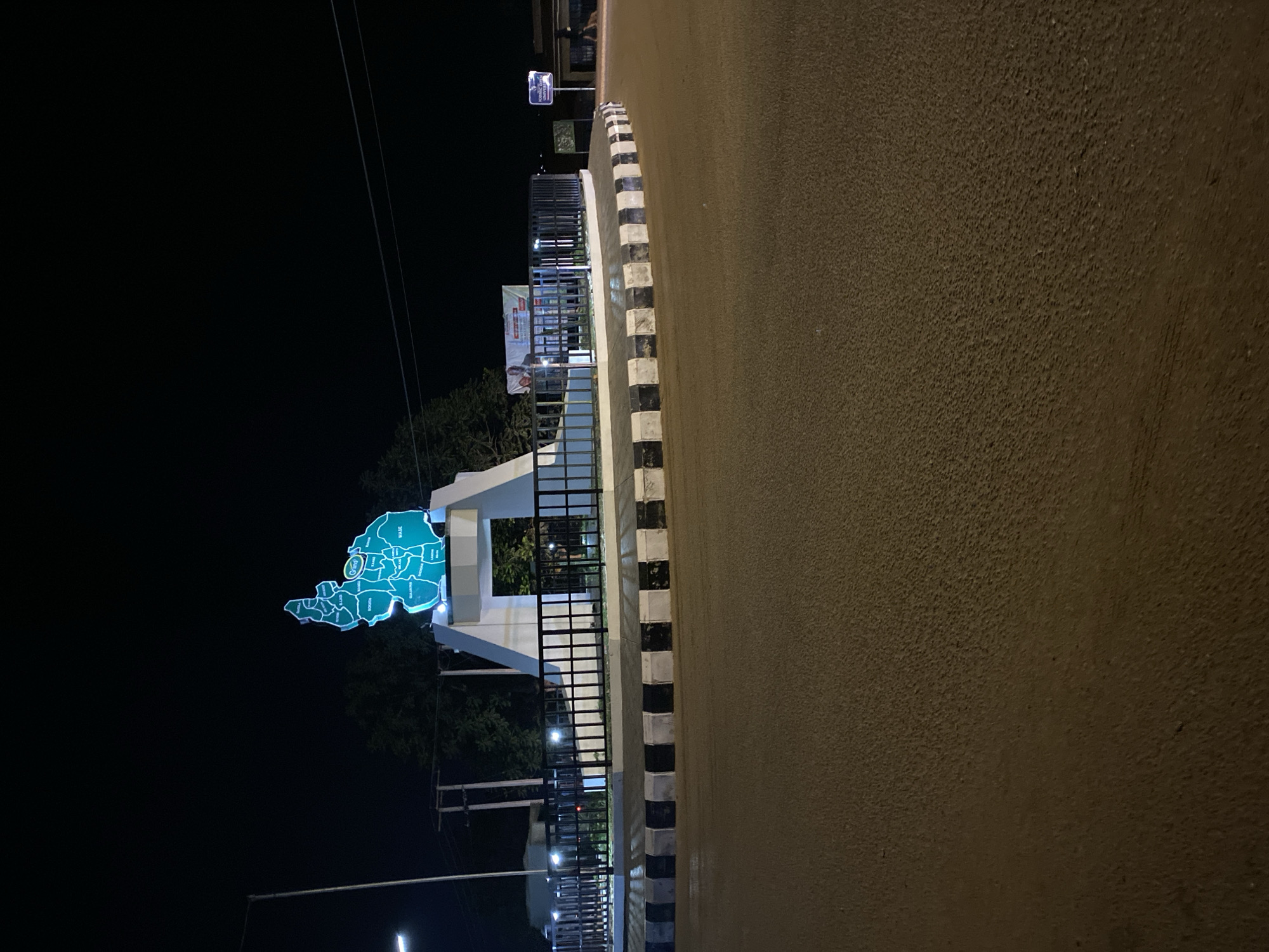 At night, a brightly lit sign in the shape of a map stands near a street in Bukuru, Nigeria. The map is a blue outline with white regions delineating areas labeled "Wase," "Mangu," "Jos," "Bassa," "Riyom," "Bokos," and "Pankshin." The map is mounted on a white architectural structure with black metal fencing. A road with black and white striped curbing is in the foreground. Street lights illuminate the area, and trees are visible behind the sign. Power lines and a billboard are visible in the background.