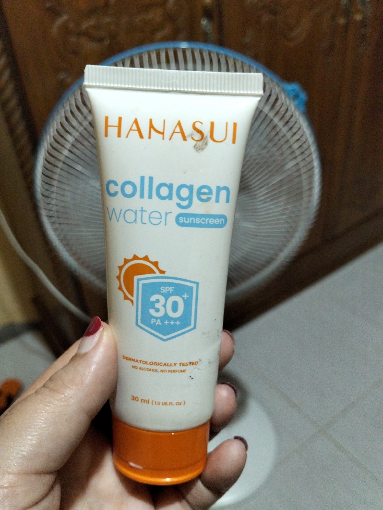 An over-the-shoulder, slightly high-angle shot shows a person's hand holding a tube of Hanasui Collagen Water Sunscreen. The tube is white with orange and blue text and graphics, featuring an orange sun icon and a blue shield with "SPF 30+ PA+++" printed inside. The tube is partially obscured by the hand and a blurred background of what appears to be a standing fan with a blue casing. The fan is positioned in front of a wooden surface, possibly furniture, and a light-colored floor is visible in the lower right corner. The lighting suggests an indoor setting.