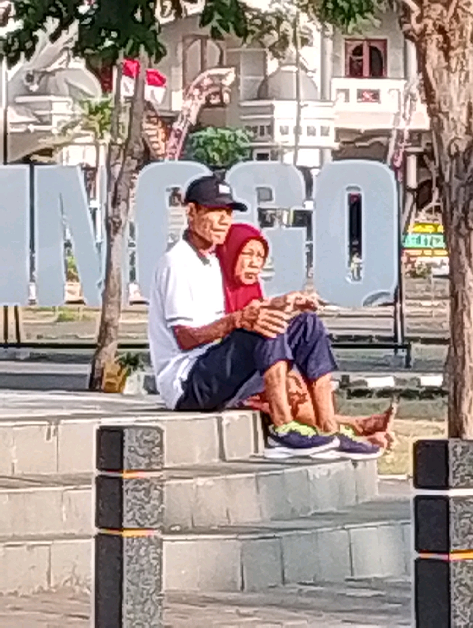 The image shows an elderly couple sitting on a tiered stone structure in a public area. The man, on the left, is wearing a white polo shirt, dark pants, and a black baseball cap. The woman, on the right, is wearing a red hijab and dark pants. They are seated side-by-side, with the man's arm around the woman. Their feet are visible, and they are both wearing casual shoes. In the background, large, grey letters are visible, which are part of a sign. Behind the sign, there are buildings with architectural details, and trees. The foreground shows more of the tiered stone structure and a decorative bollard. This scene is located in front of the Probolinggo City Hall in Probolinggo, Indonesia, as indicated by the prominent display of the city's name.