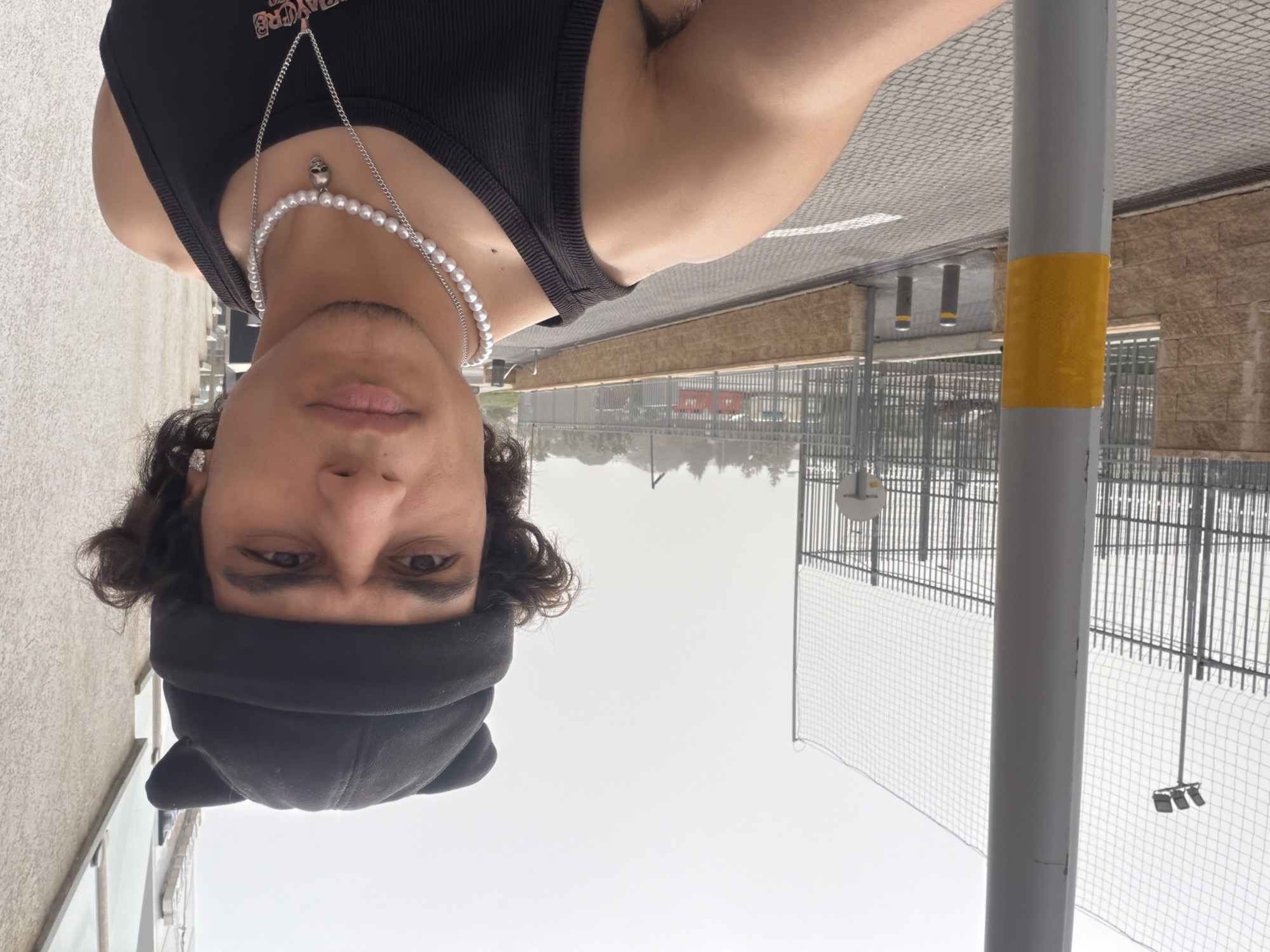 A young person, likely male, is captured in a vertical eye-level shot, posing outdoors in the Hof HaCarmel Regional Council, Israel. The subject looks directly at the camera with a neutral expression. They are wearing a black ribbed tank top with partially visible stylized text that appears to be "PLAYBOY" printed on it. Adorning their neck is a pearl necklace and a separate silver chain featuring a skull-shaped charm. A black beanie with subtle ear-like protrusions covers their forehead, and dark, curly hair frames their face. Light facial stubble is also visible.
The background reveals an overcast sky, indicating a daytime shot with soft, diffused lighting. To the right of the subject, a prominent grey pole with a yellow band stands vertically. Beyond this pole and extending to the right, a tall metal fence and a mesh net, possibly part of a sports court or enclosed facility, are visible. In the distance, light-colored buildings with textured walls are present. The ground beneath the subject appears to be paved with patterned tiles or cobblestones. The scene depicts a casual outdoor setting within a fenced area.