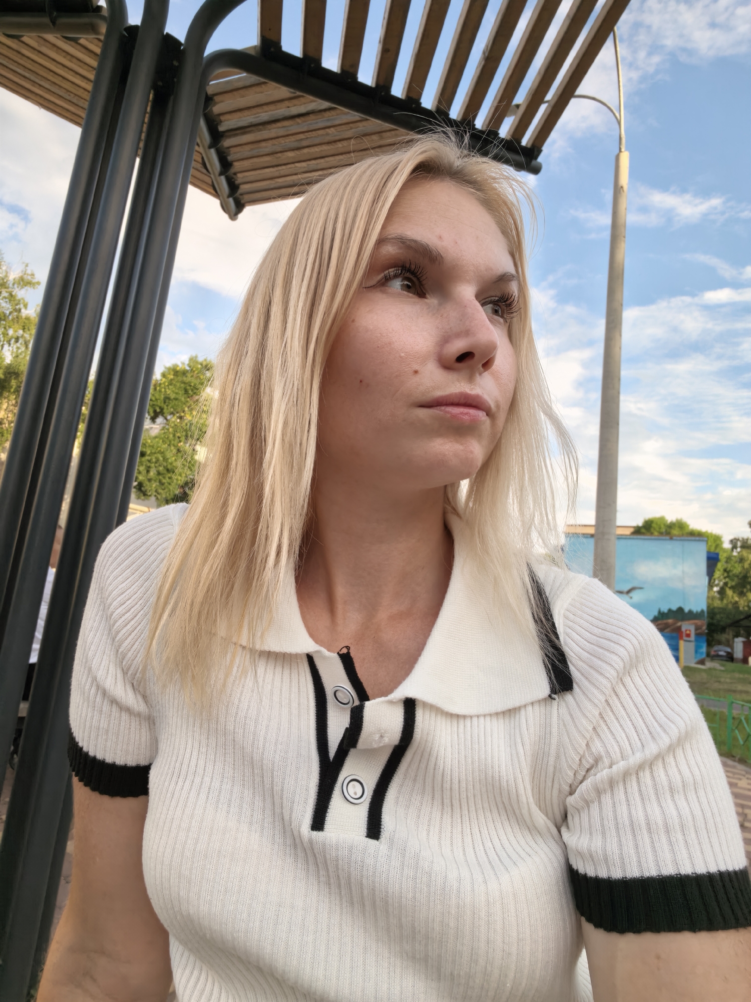 The image shows a young Caucasian woman with blonde hair and light skin, wearing a white short-sleeved polo shirt with black trim and buttons, sitting outdoors under a wooden canopy. She is looking to the right of the frame, with a neutral expression. In the background, there is a blue sky with scattered clouds, trees, a street lamp, and a building with a mural on its side. The setting appears to be a park or public area, possibly in the city of Sterlitamak, Russia, given the context. There are metal poles supporting the canopy in the foreground.