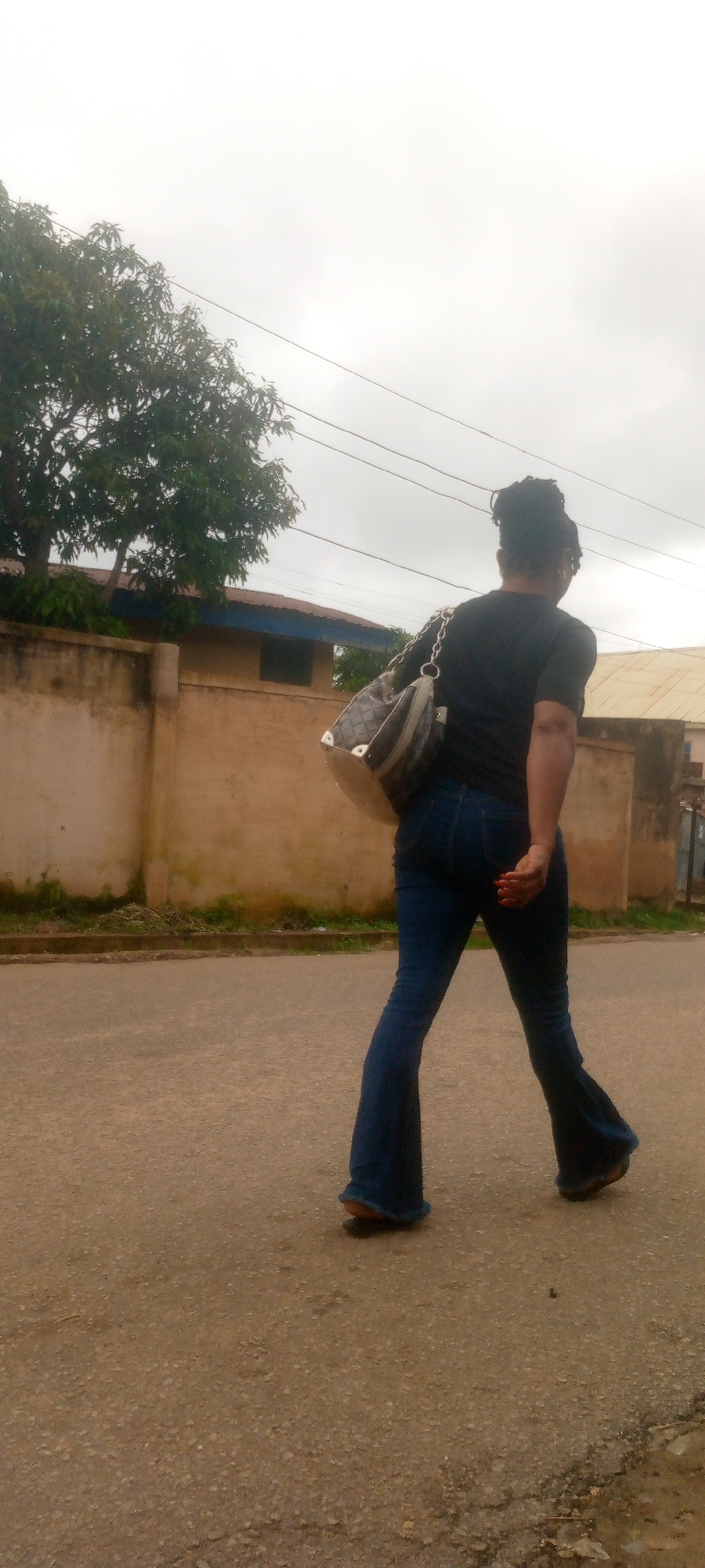 A medium shot captures a Black woman from behind, walking away from the camera on a paved road. She is wearing a black t-shirt and blue bell-bottom jeans. A patterned shoulder bag with a chain strap is slung over her left shoulder. Her hair is styled in an updo. In the background, a textured tan wall runs horizontally, with a tree and the corner of a building visible beyond it. The sky is overcast and light. The setting appears to be a residential or semi-urban area, with the road being the primary focus of the foreground.
