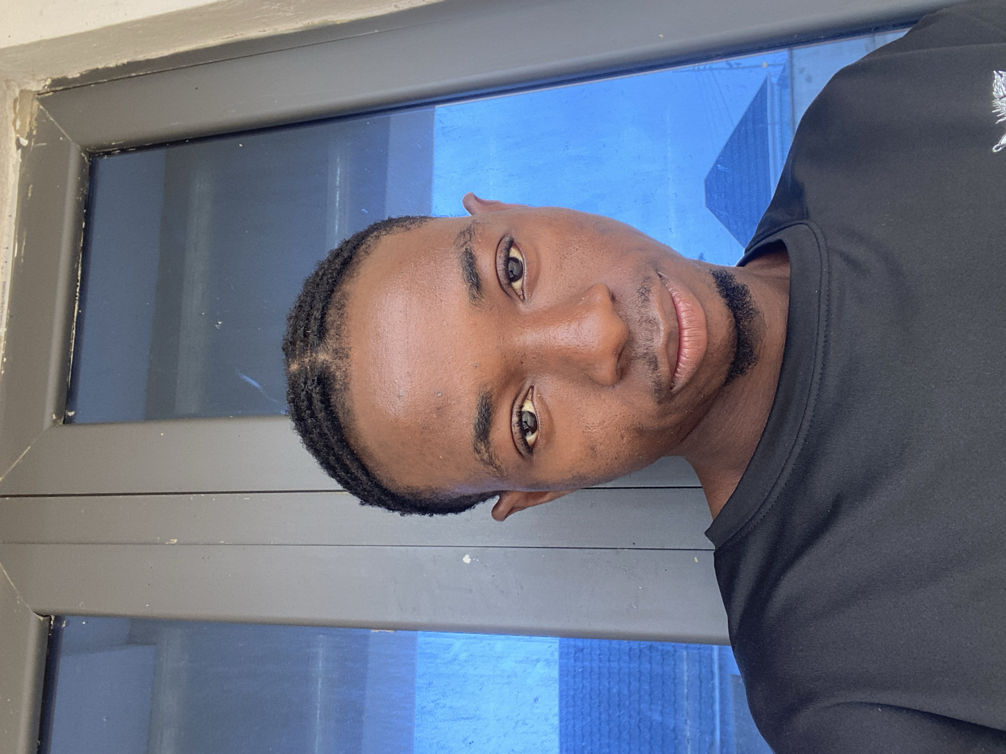 A young Black man with braided hair and a goatee is captured in a head and shoulders selfie. He is wearing a black t-shirt and is standing in front of a large window. The window has blue panes and a grey frame. The background visible through the window appears to be an outdoor scene with some structures that could be buildings. The man's skin tone is dark brown, and his eyes are dark. He has a slight smile. The lighting suggests an outdoor or well-lit indoor environment. The image does not provide specific geographical markers that would allow for identification of the city of Jos, Nigeria.