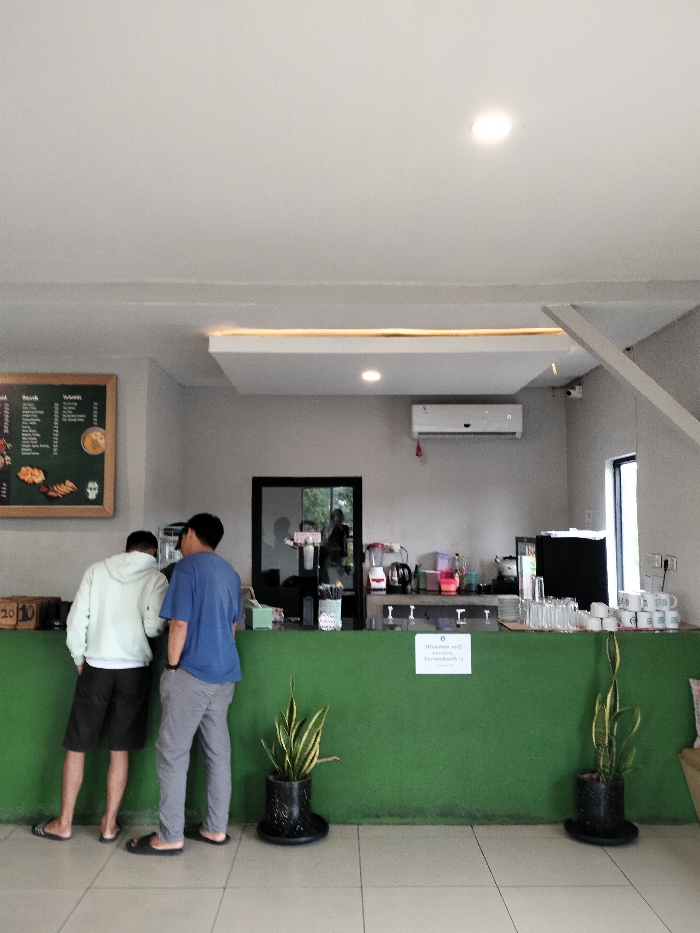 This image depicts an indoor scene at a casual self-service establishment, likely a cafe or restaurant, in Dramaga, Indonesia. Two men are standing with their backs to the camera at a prominent green counter. The man on the left wears a light-colored hoodie and dark shorts, while the man on the right is in a blue t-shirt and grey pants; both are wearing flip-flops. They appear to be ordering or waiting for items at the counter.
Behind the green counter, the background features a light grey wall equipped with an air conditioning unit and a security camera. Various items typically found in a service area are visible, including blenders, kettles, stacks of clean glasses, and white mugs. A dark-framed doorway or window in the back wall reflects a blurred figure. On the left wall, a dark menu board lists food and drink options, with headings like "Food" and "Drink," though specific items are mostly illegible.
On the green counter, a small white sign clearly states "Minuman self service" (Drink self-service) and "Terima Kasih" (Thank You), indicating a self-service beverage station. Two potted snake plants sit on the floor in front of the counter, adding natural decor. The ceiling is white with recessed lighting and a decorative dropped panel featuring strip lighting. The floor is light-colored tiled, and the space is brightly lit, suggesting it is daytime. The overall setting is functional and relaxed.