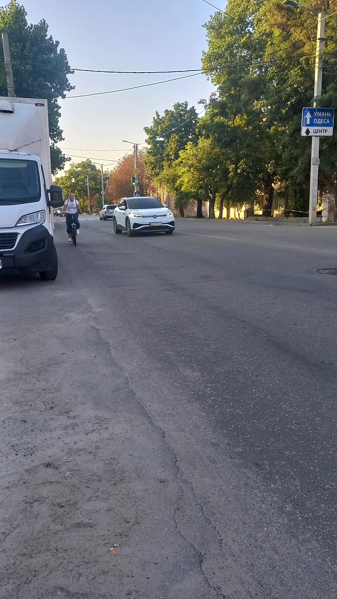The image depicts a street scene in Kropyvnytskyi, Ukraine. In the foreground, a white van is parked on the left side of the road, partially obscuring the view of a person riding a bicycle in the middle lane. The cyclist, wearing a white t-shirt and dark trousers, is facing forward and appears to be moving. A white electric car is traveling in the right lane ahead of the cyclist. A street sign with blue lettering and an arrow pointing upwards is visible on the right side of the road, indicating directions to "UMAN ODESSA" and "CENTER". Trees line the right side of the street, with foliage casting shadows on the road. The sky is clear and blue, suggesting fair weather. The road surface appears to be asphalt with some visible cracks and wear. The overall scene suggests a typical urban street during daytime.
