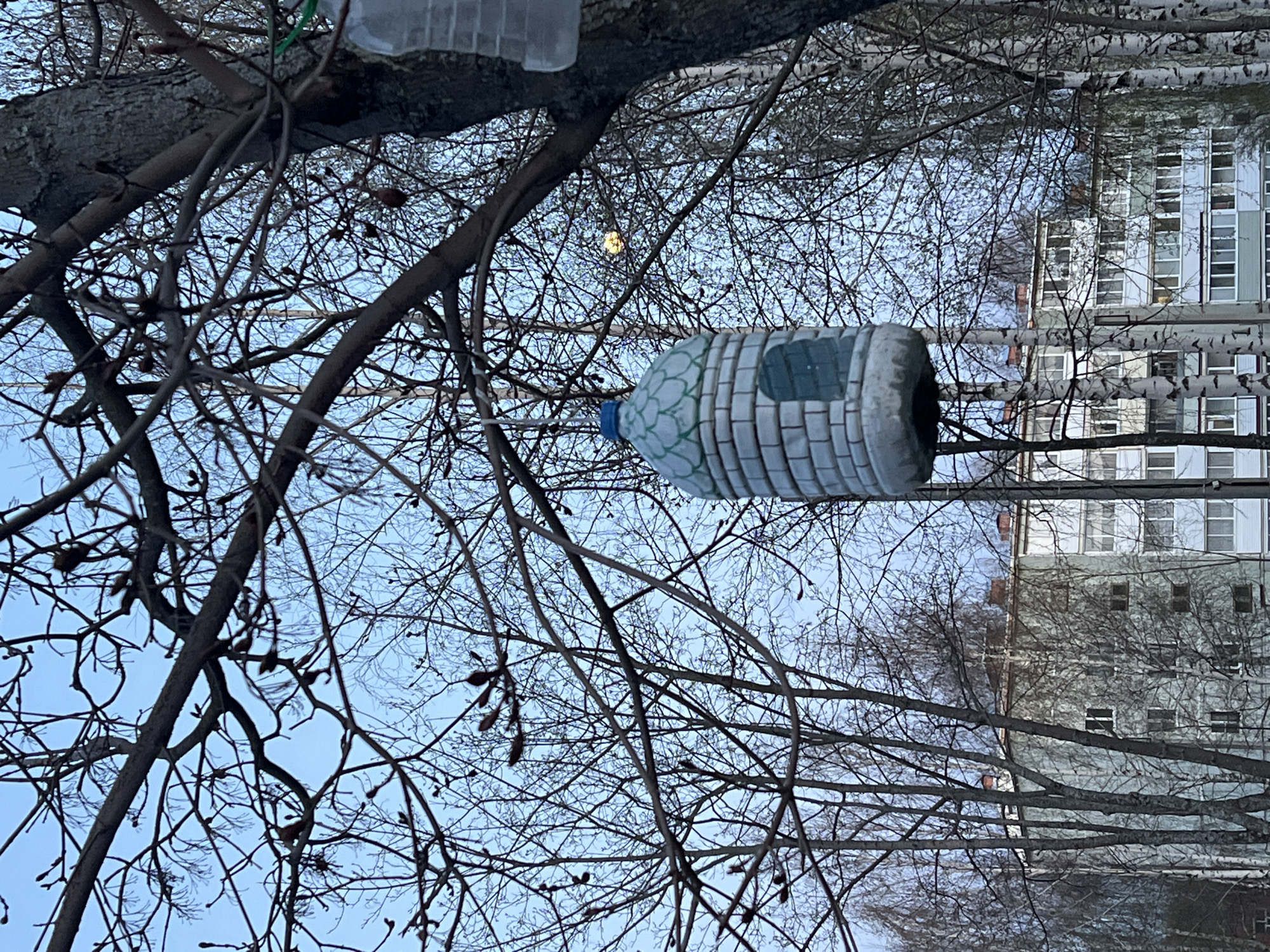 A repurposed plastic bottle bird feeder hangs from a bare tree branch in Noviy Urengoy, Russia. The bottle, painted to resemble a brick structure with a dark window, is suspended against a backdrop of a multi-story building with white-framed windows. The sky is a pale blue, and the bare branches of other trees fill the space, obscuring parts of the building. Another bottle is visible in the upper part of the picture.
