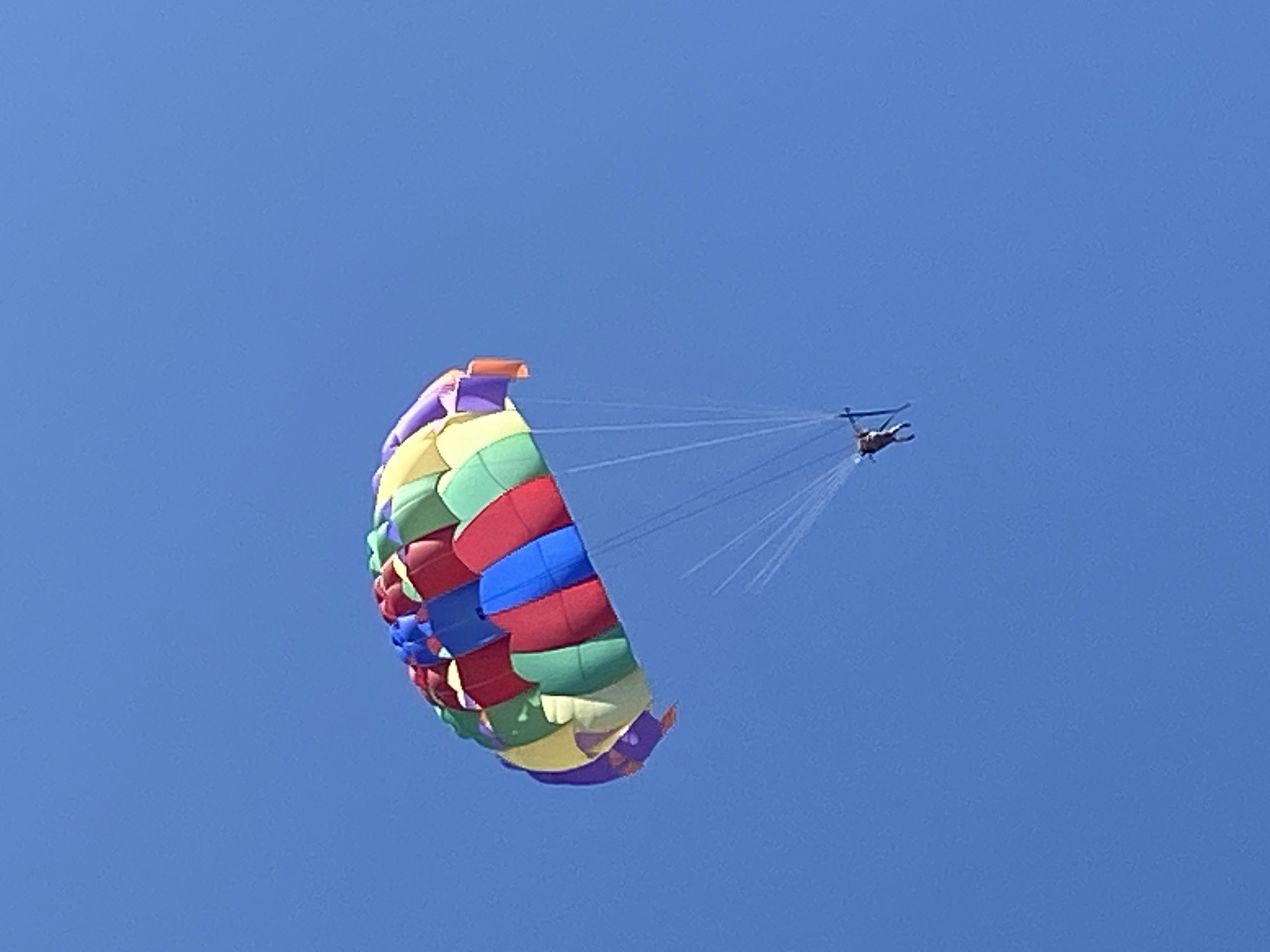 A parasail in flight against a clear blue sky. The parasail is multicolored and domed, with visible tow ropes connecting it to a distant figure or object, possibly a boat. The specific location in Prijevor, Montenegro cannot be determined from the visual information.