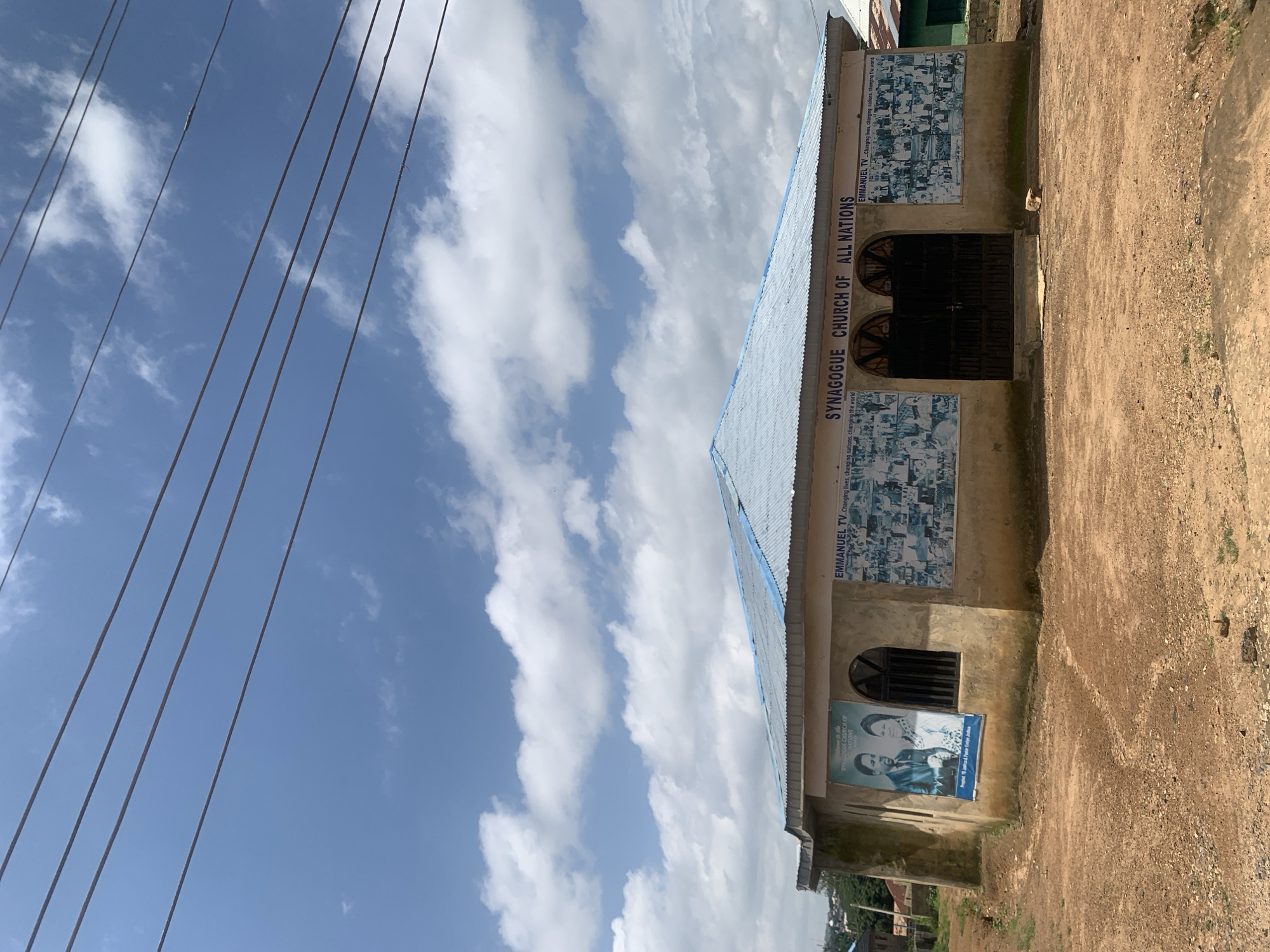 A medium shot captures the Synagogue Church of All Nations in Rafin Par, Nigeria, under a partly cloudy sky. The church, a concrete structure with a corrugated metal roof painted blue, has a tan exterior. The front of the building is adorned with posters. A large poster on the left displays an image of a man and a woman, with text below that reads "Prophet T.B. Joshua & Pastor Evelyn Joshua." To the right of this, above the entrance, is a sign that reads "SYNAGOGUE CHURCH OF ALL NATIONS." Flanking the entrance are two large posters filled with smaller images of crowds of people. The entrance itself features two dark metal gates. The ground in front of the church is unpaved, composed of reddish-brown dirt and gravel, with some scattered stones. To the right of the church, a sliver of a green corrugated metal roof is visible. Power lines are strung across the upper portion of the frame. The overall scene suggests a rural or semi-urban setting.