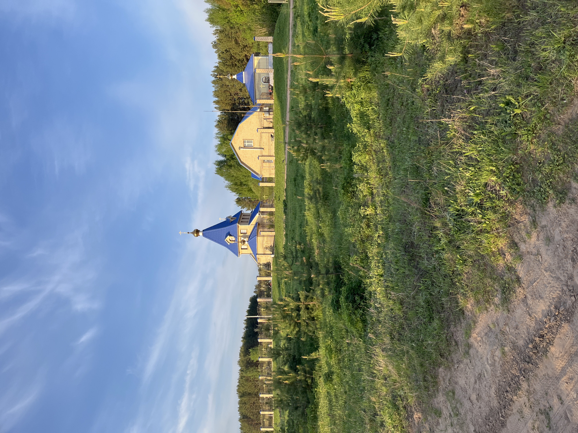 The image shows a religious building, likely an Orthodox church, in Chaikovsky, Russia. The building has a blue roof and a golden dome topped with a cross. There are other similar buildings nearby. The area surrounding the building is grassy and has a dirt path. The sky is blue with light clouds. The scene appears to be taken during the day in a rural or suburban area.