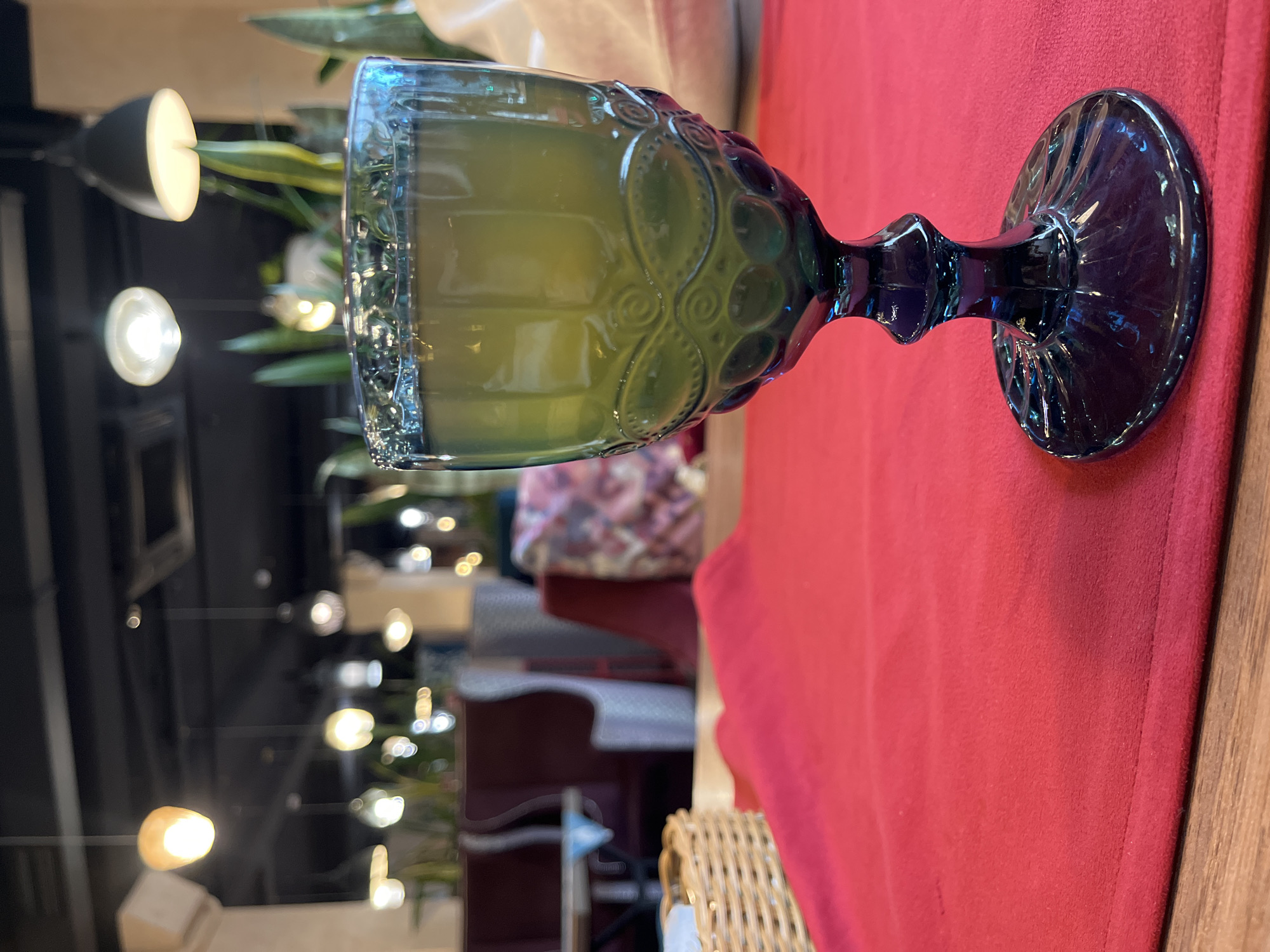 The image prominently features a decorative, goblet-style glass filled with a yellowish-green liquid, set on a vibrant red tablecloth. The glass itself is a deep blue-green, possibly made of colored glass, and is adorned with intricate embossed patterns around its bowl and a faceted stem. Its base is also patterned.
The red tablecloth occupies the majority of the foreground and midground, with a strip of wooden table surface visible along the right edge. The background is softly blurred, creating a bokeh effect from numerous artificial light sources. These lights appear as glowing circles of various sizes, indicating an indoor setting like a restaurant, cafe, or bar, likely during the evening. Partially visible in the blurry background are some green potted plants, adding a touch of nature to the interior. Other indistinct shapes suggest tables, chairs, or architectural elements of the establishment. A wicker texture, possibly from a basket or chair, is also faintly discernible in one of the blurred areas.
No people are visible in the frame, and there is no discernible text. The scene conveys a calm, inviting atmosphere, characteristic of a dining or social establishment in Izhevsk, Russia.
