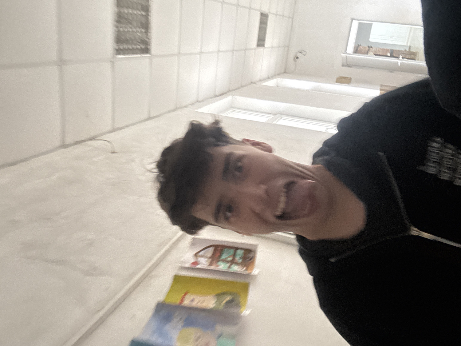 This image features a young male, likely a teenager or young adult, captured from a low-angle perspective in an indoor setting in Troyan, Bulgaria. He is in the foreground, with his head tilted and a playful expression on his face, sticking out his tongue and showing his teeth. He wears a dark, possibly black, zipped hoodie, which has some illegible white text or design on the right shoulder area.
The background consists of light-colored, possibly white or off-white, walls displaying a grid-like pattern, which could be tiles or panels. High up on these walls are windows, letting in natural daylight, indicating it is daytime. Below the person, on what appears to be a counter or shelf, several flat objects resembling drawings or paintings are stacked, suggesting an area where art is displayed or created. The overall mood of the image is casual and humorous.