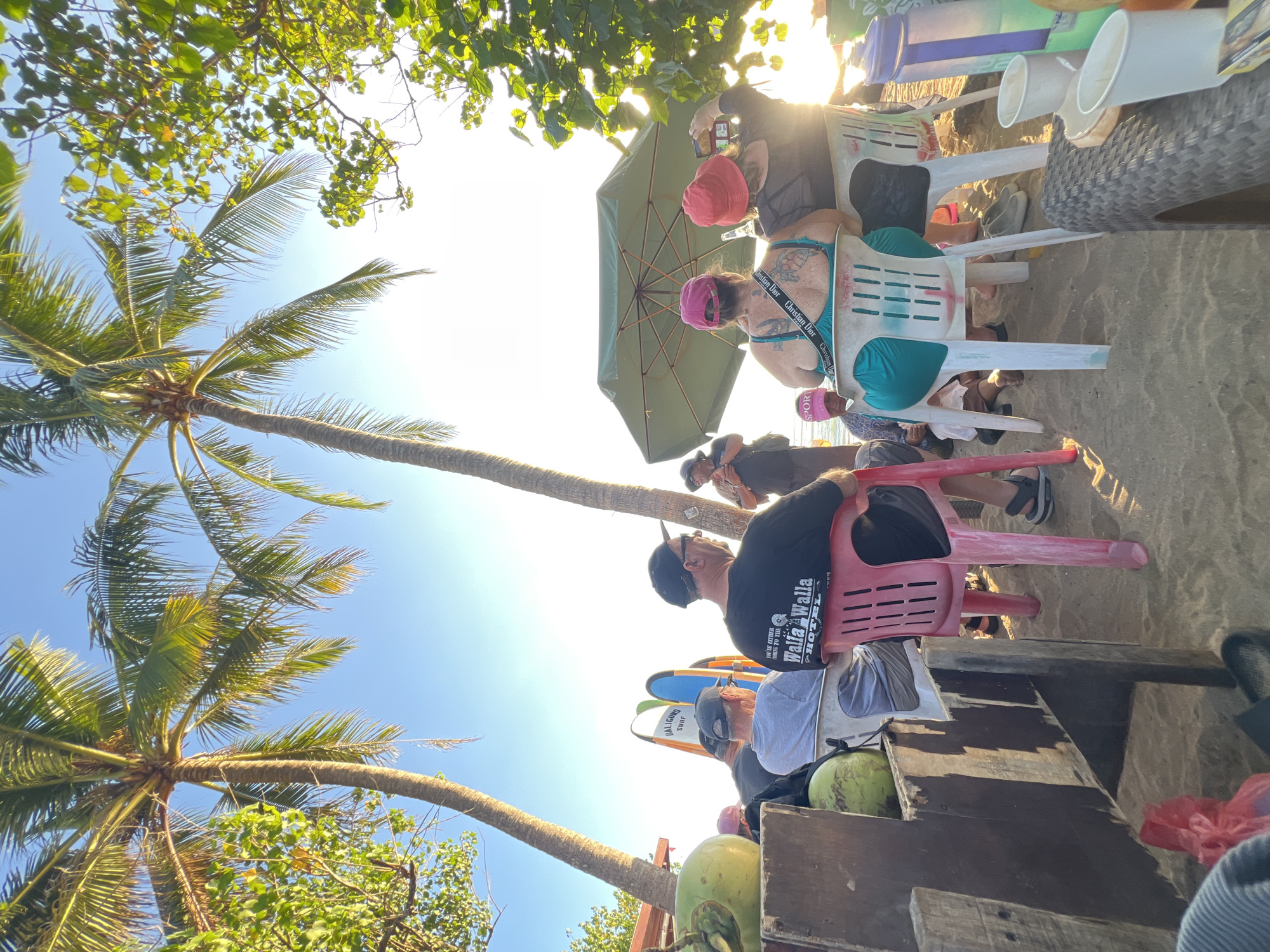 This is a photograph taken at a beachside venue in Kuta, Indonesia, during a sunny day. The scene is composed of several individuals seated on chairs, with two prominent palm trees overhead and lush green foliage to the right. In the foreground, a wooden structure with coconuts rests on the sand. The individuals are casually dressed, and some appear to be engaged in conversation or enjoying the scenery. A person in the center appears to be playing a musical instrument, while another is taking a photograph. Surfboards are visible leaning against a wall in the background. The overall ambiance suggests a relaxed gathering at a tropical beach.