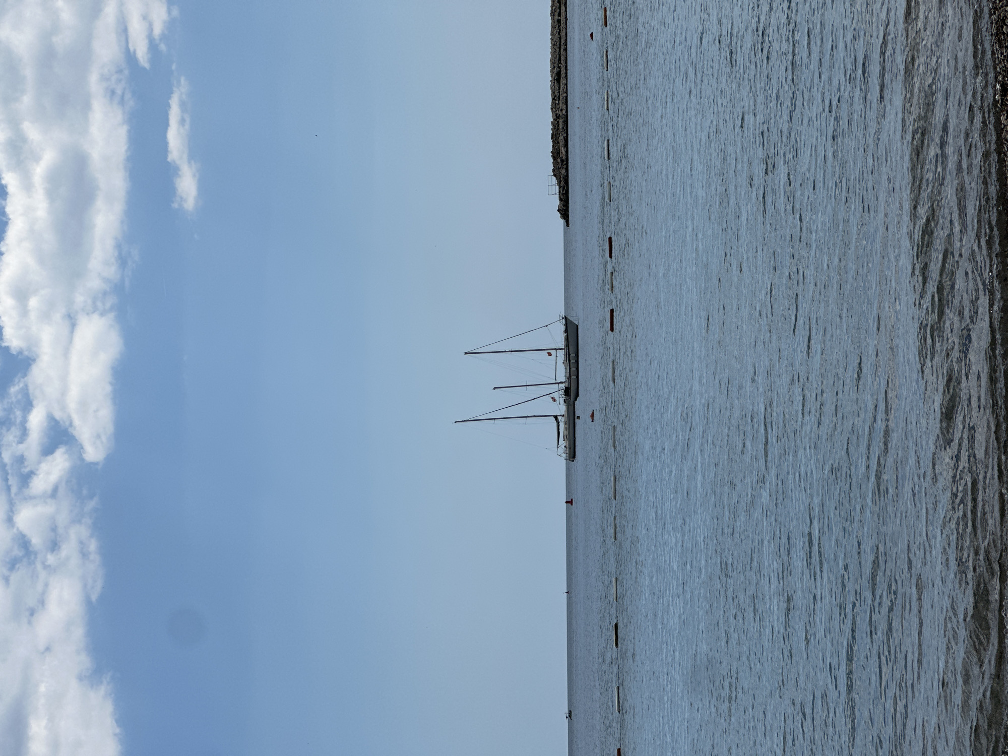 The image depicts a scene at the sea near Budva, Montenegro. The sky is partly cloudy with a clear blue hue. Two sailboats are anchored or moored in the sea. A breakwater is visible near the top edge of the image. The sea is calm and reflects the sky. Buoys mark a swimming area. The water appears to be shallow in the immediate foreground. The angle and proximity to the sea suggest the photograph was taken near the beach.