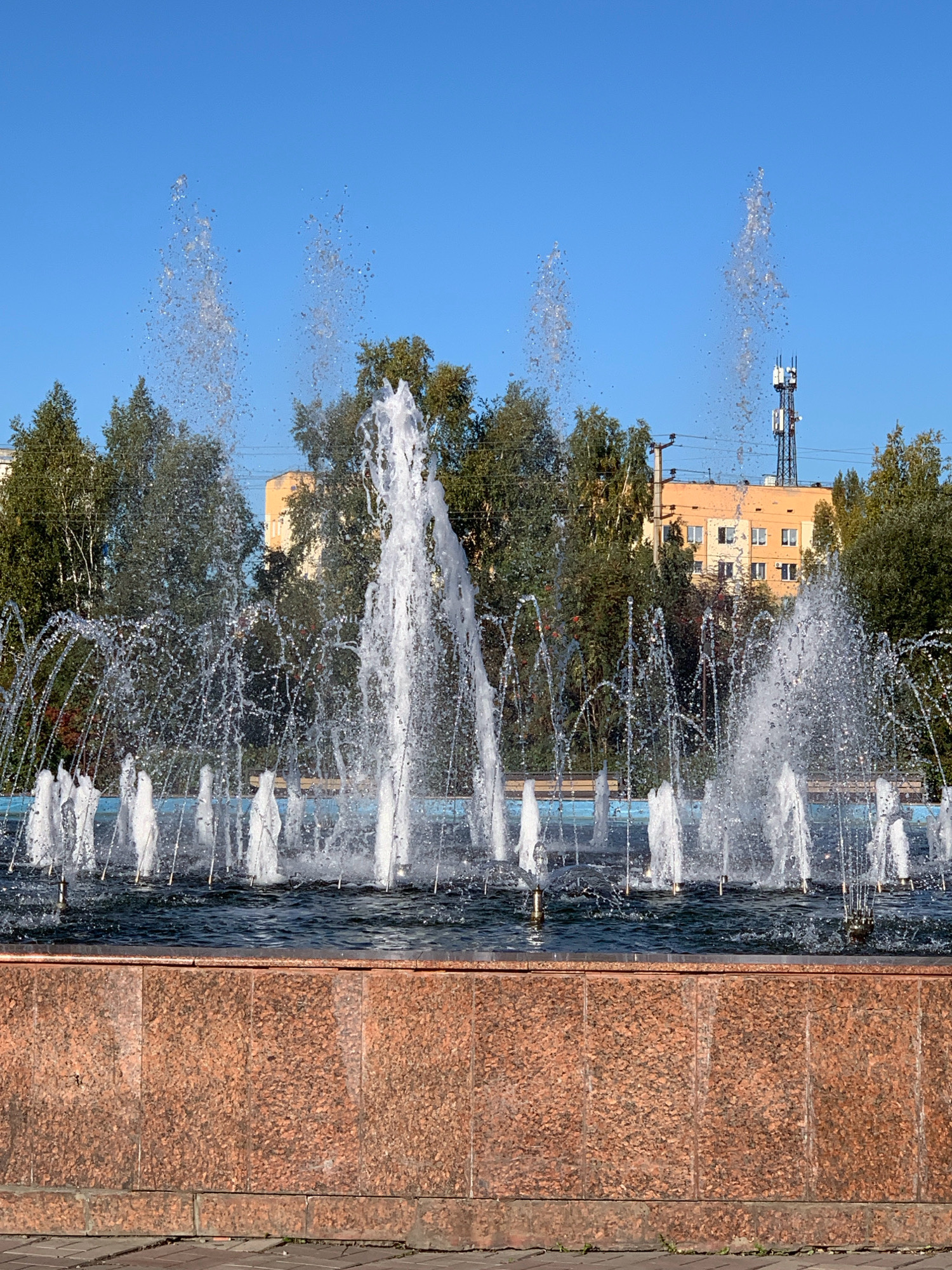 The image shows a public fountain in the city of Prokopyevsk, Russia. The fountain features multiple jets of water spraying upwards into a clear blue sky. The water is creating a dynamic spray and mist. In the background, there are several green trees and some buildings, including a light orange apartment building and a cell tower. The fountain's basin is surrounded by a reddish-brown stone structure. The foreground shows a paved area with bricks. There are no people visible in the image.