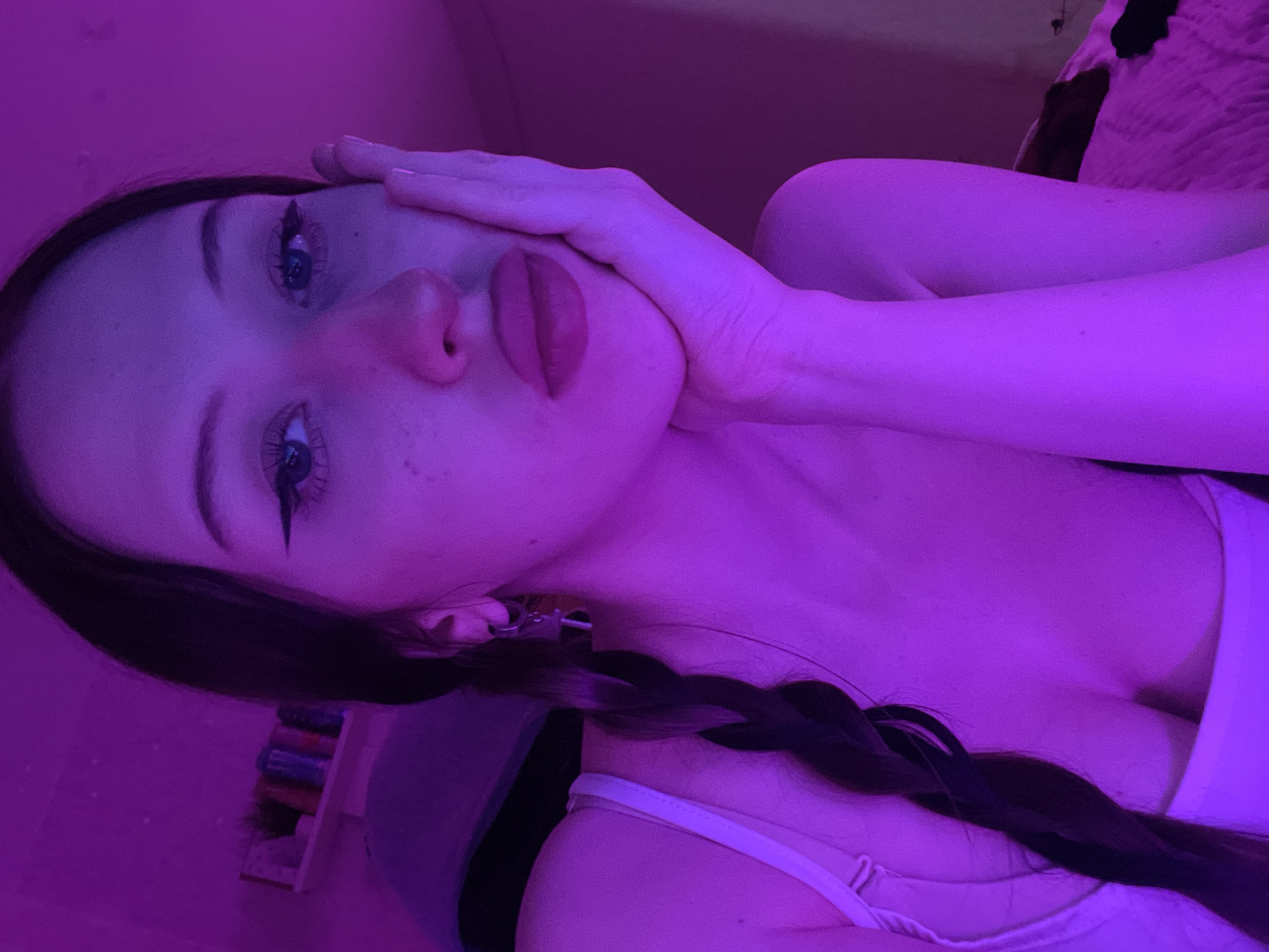 The image was captured indoors in the city of Votkinsk, Russia. It features a young woman posed with her chin resting on her right hand, looking directly at the viewer. She has long, dark hair styled in a braid over her left shoulder. Her eyes are accentuated with winged eyeliner, and her lips appear full. A small key earring is visible on her left ear. She is wearing a white garment, possibly a camisole or bra, partially visible on her torso. The entire scene is illuminated by a strong, uniform magenta or purple light source, creating a vibrant, monochromatic atmosphere. In the background to the left, a shelf holds several indistinct containers. To the right, a textured surface, likely a bed, is visible.