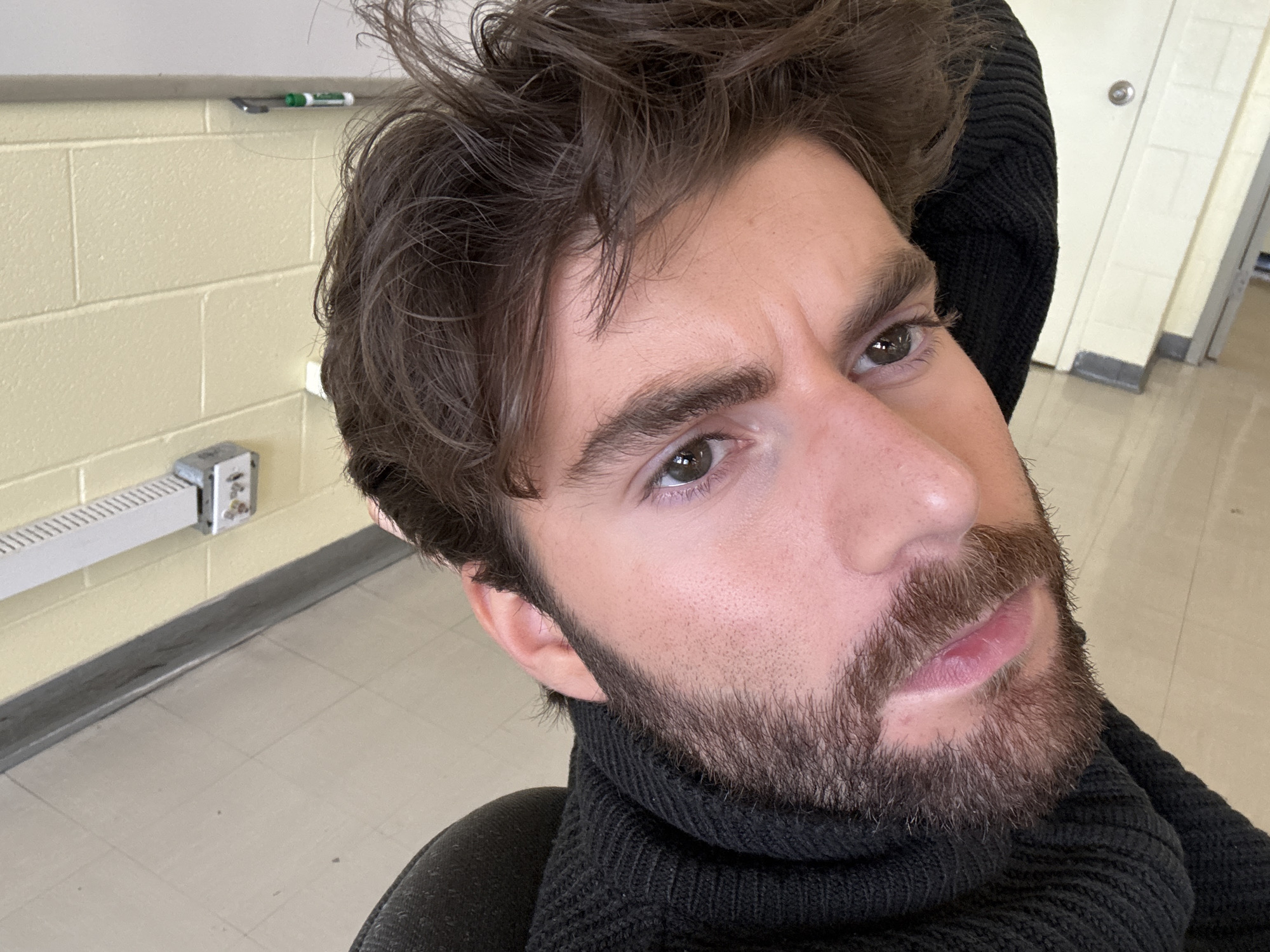 A close-up selfie of a man with brown hair and a beard, wearing a black turtleneck sweater. The background shows a light beige brick wall with a white whiteboard and a green marker resting on it. Below the wall is a white radiator. The floor is tiled in a neutral pattern. The location appears to be an interior room with institutional characteristics, possibly a classroom or office setting. The exact location within Montreal, Canada is impossible to determine from the image.