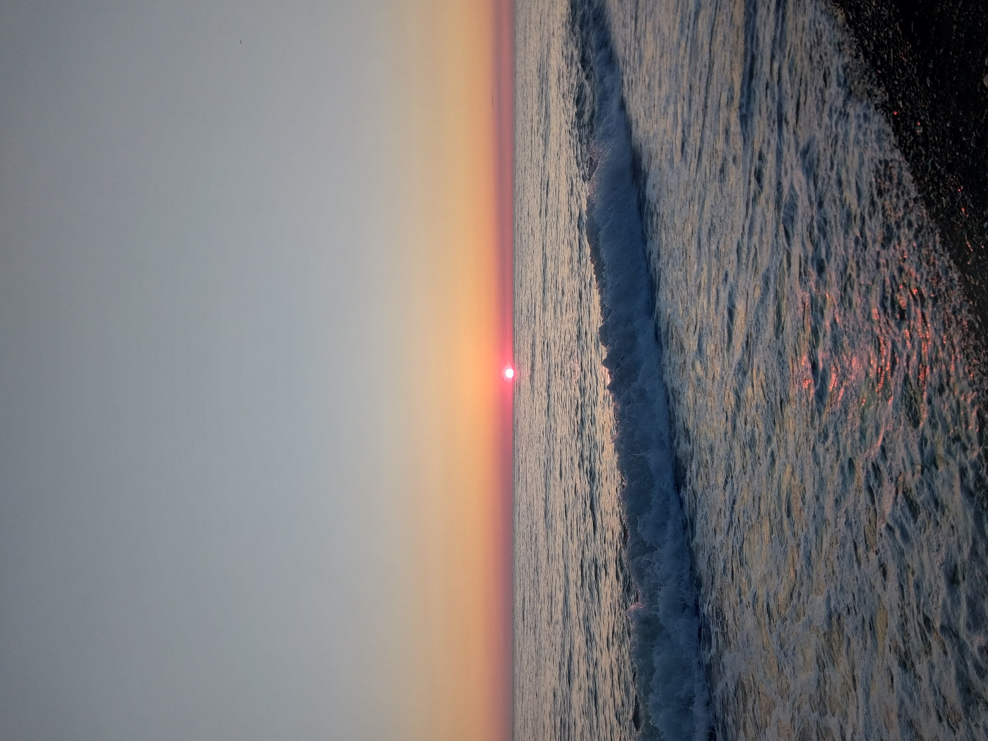 The image displays a serene coastal landscape captured during either sunrise or sunset, centered on the horizon line of a body of water, likely the sea or ocean, with the provided context suggesting Sochi, Russia.
The primary subject is the sun, appearing as a vivid, bright red-orange orb resting directly on the horizon. Above the horizon, the sky exhibits a soft gradient, transitioning from warm hues of orange and pink near the sun to a cooler, pale blue-grey further up and to the left. A faint, small dark speck, possibly a distant bird, can be observed in the upper left portion of the sky.
To the right of the horizon, the ocean dominates, showcasing active waves breaking and moving towards the shore. The water's surface is rich with texture, displaying ripples, foam, and the distinctive white crests of an incoming wave. The warm light from the low-lying sun casts subtle reflections on the water, adding hints of red and orange to the otherwise cool, dark blue and grey tones of the sea.
The extreme right edge of the frame reveals a dark, rugged shoreline, possibly composed of pebbles or rocks, where the ocean waves meet the land. The scene is devoid of any human presence or man-made objects, reinforcing a sense of untouched natural beauty and tranquility. The quality of light strongly indicates the early morning or late evening hours. There is no visible text in the image.