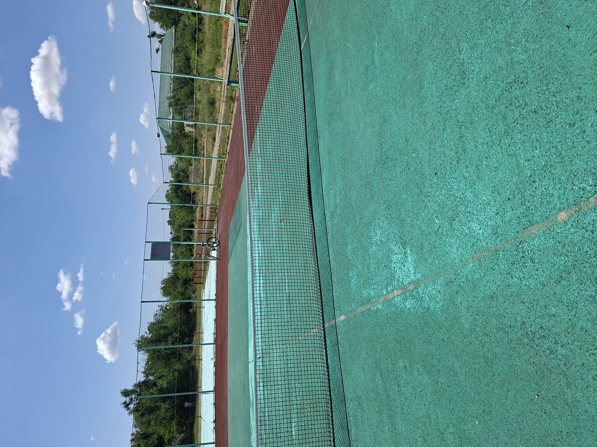 An outdoor tennis court with a green surface and a red border is shown. A net divides the court. In the background, there is another tennis court with a blue surface, fenced by a green metal mesh. A bicycle is parked next to the fence. Trees and a building with a green roof are visible beyond the fence. The sky is blue with scattered white clouds. The scene appears to be located in Gorkovskiy, Russia.