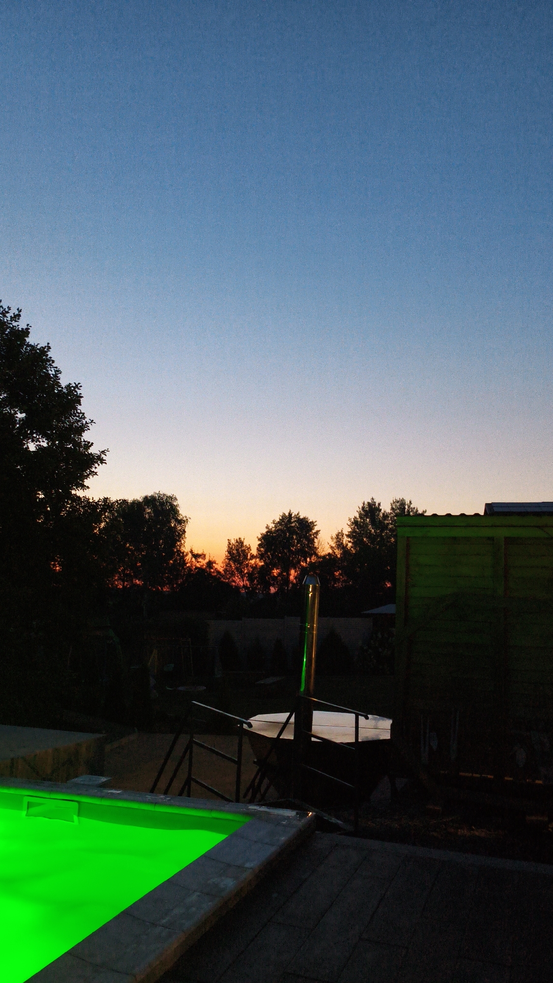 In Bezliudivka, Ukraine, a swimming pool with bright green lighting is visible in the foreground, situated next to a paved area. Beyond the pool, a metal staircase leads up to a structure. In the middle ground, trees are silhouetted against a sunset sky, with an orange glow near the horizon. A tall, metallic chimney with green lighting rises vertically. In the background, a wooden shed with a green roof is present. The sky transitions from a clear blue at the top to a warm gradient of orange and pink near the horizon, indicating dusk.