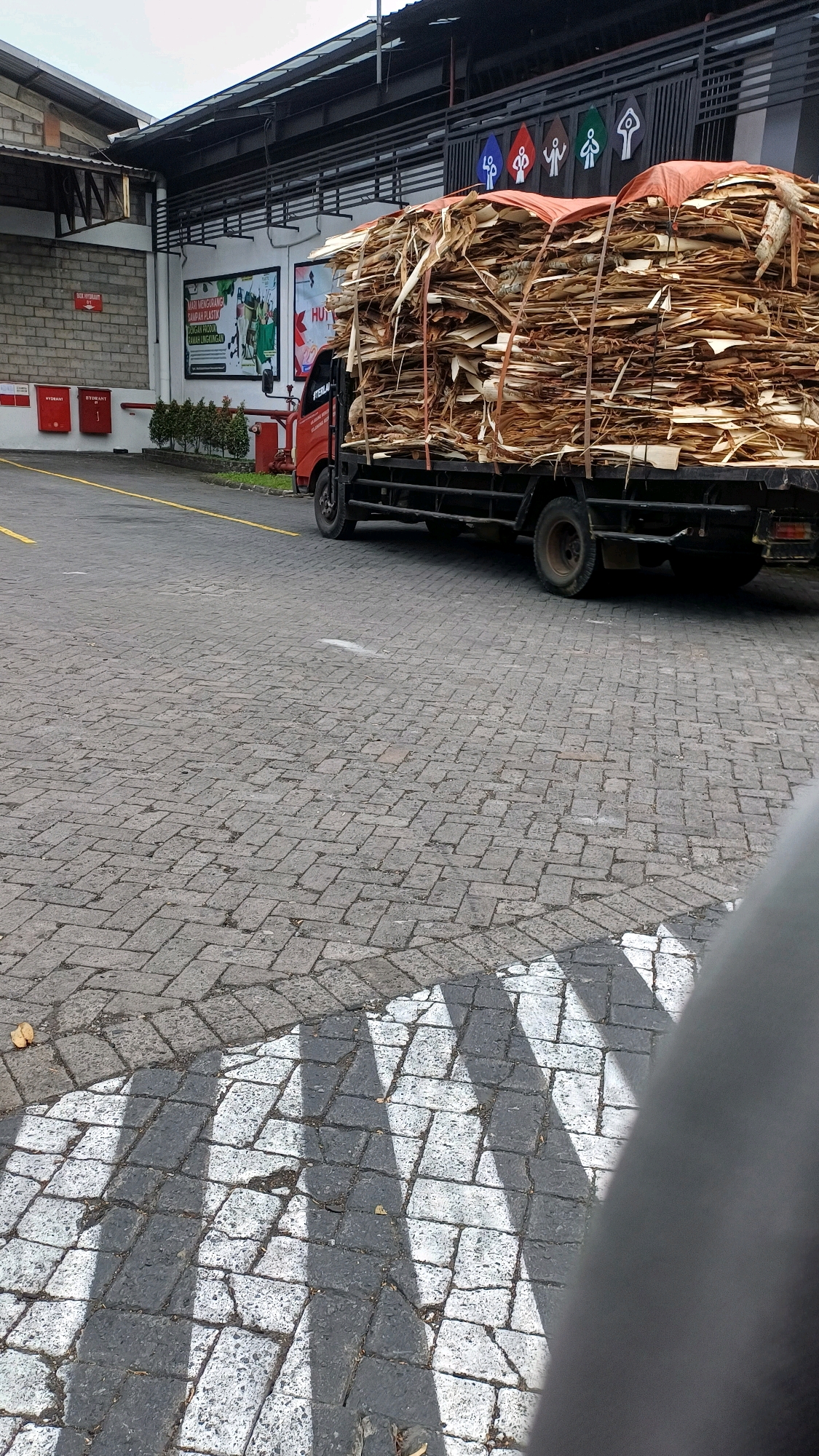 A truck laden with a large load of dried leaves or wood is parked on a cobblestone street in the city of Kranggan, Indonesia. The truck's cargo is covered with a tarp and secured with ropes. In the background, there is a building with a red and white sign that reads "UTAMA" and another sign with the text "HUT" and an image of a hand holding a leaf. There are also decorative figures on the building's facade. A crosswalk with black and white stripes is visible on the street in the foreground. The overall scene depicts an industrial or commercial area.