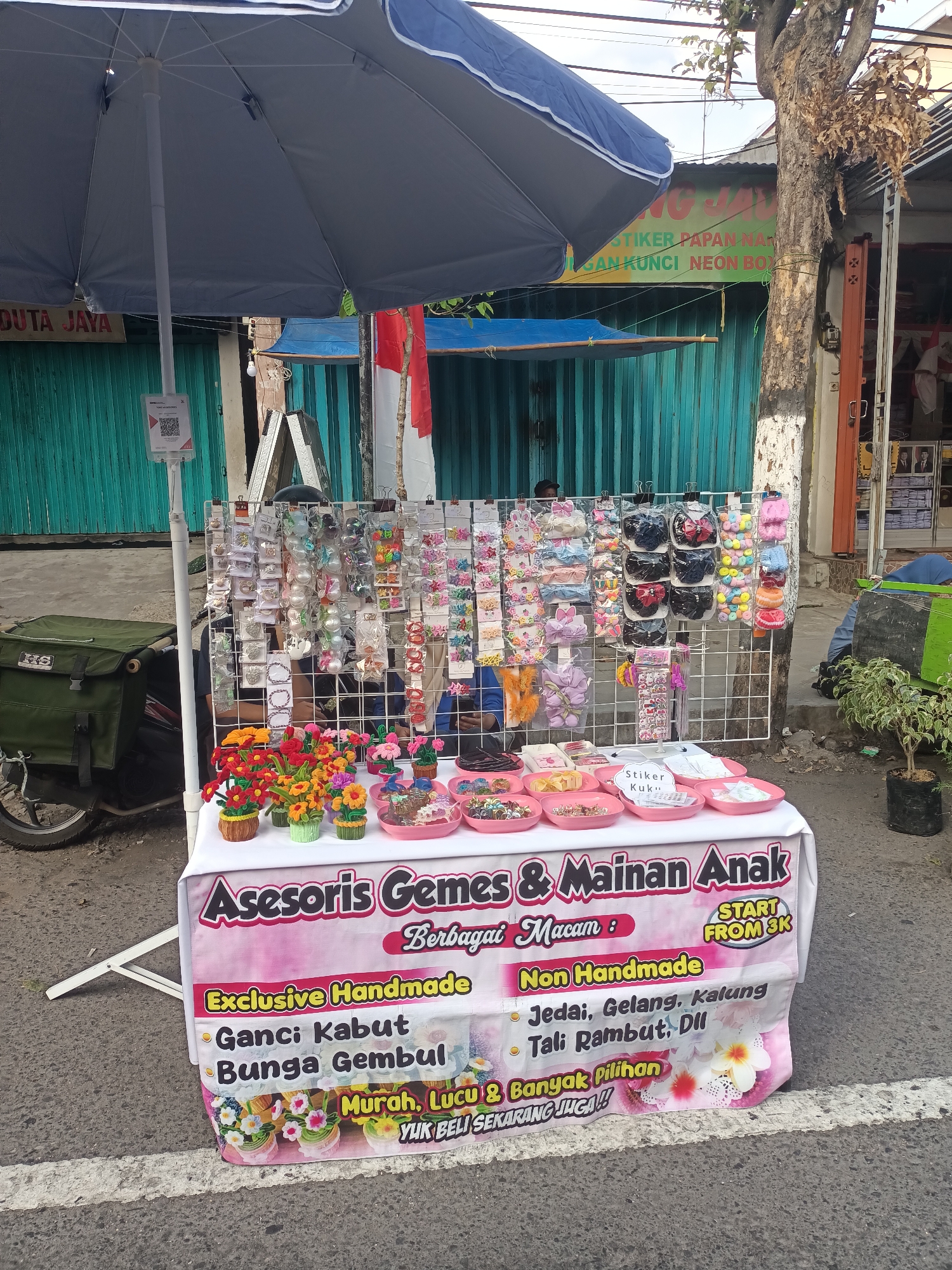 The image shows a street stall selling accessories and toys for children in Pare, Kediri, Indonesia. The stall is set up under a large blue umbrella and features a variety of items, including hair clips, hair ties, bracelets, and necklaces. The stall also sells handmade items such as flower bouquets and possibly charms. The banner at the front of the stall reads "Asesoris Gemes & Mainan Anak" which translates to "Cute Accessories & Children's Toys." Below this, it lists "Berbagai Macam" meaning "Various Types," and "Exclusive Handmade." Specific items mentioned are "Ganci Kabut Bunga Gembul" (Charm, Cloud, Chubby Flower) and "Jedai, Gelang, Kalung, Tali Rambut, dll" (Hair Clamps, Bracelets, Necklaces, Hair Ties, etc.). The banner also advertises "Murah, Lucu & Banyak Pilihan" meaning "Cheap, Cute & Lots of Choices," and a call to action "YUK BELI SEKARANG JUGA!!" which means "Let's buy now!!". The stall also displays "START FROM 3K," indicating the starting price of the items. In the background, there is a building with green metal doors, a tree, and electrical wires overhead. A motorcycle with a green bag is parked to the left of the stall. A person is partially visible behind the stall. The street is paved with asphalt.