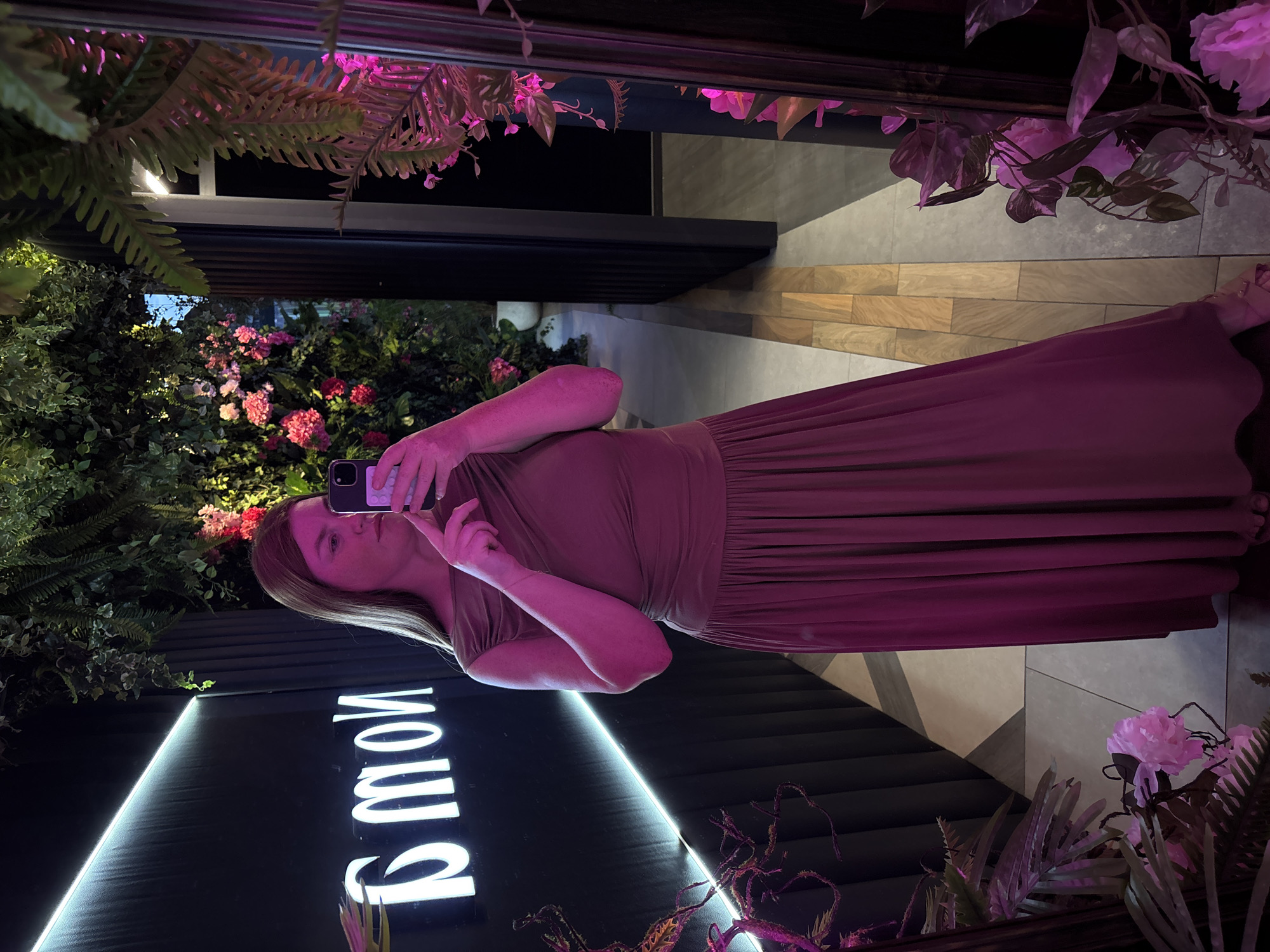 A woman takes a selfie in a mirror, her reflection showing her from the chest up. She is wearing a long, mauve dress. The background features lush green foliage, pink flowers, and a neon sign that reads "MOM" in large, white letters. The scene appears to be indoors, possibly a restaurant or lounge, with modern tile flooring and ambient purple lighting. The woman's expression is neutral as she focuses on her phone. The overall mood is somewhat dramatic and stylish, with the contrasting elements of nature and artificial light.