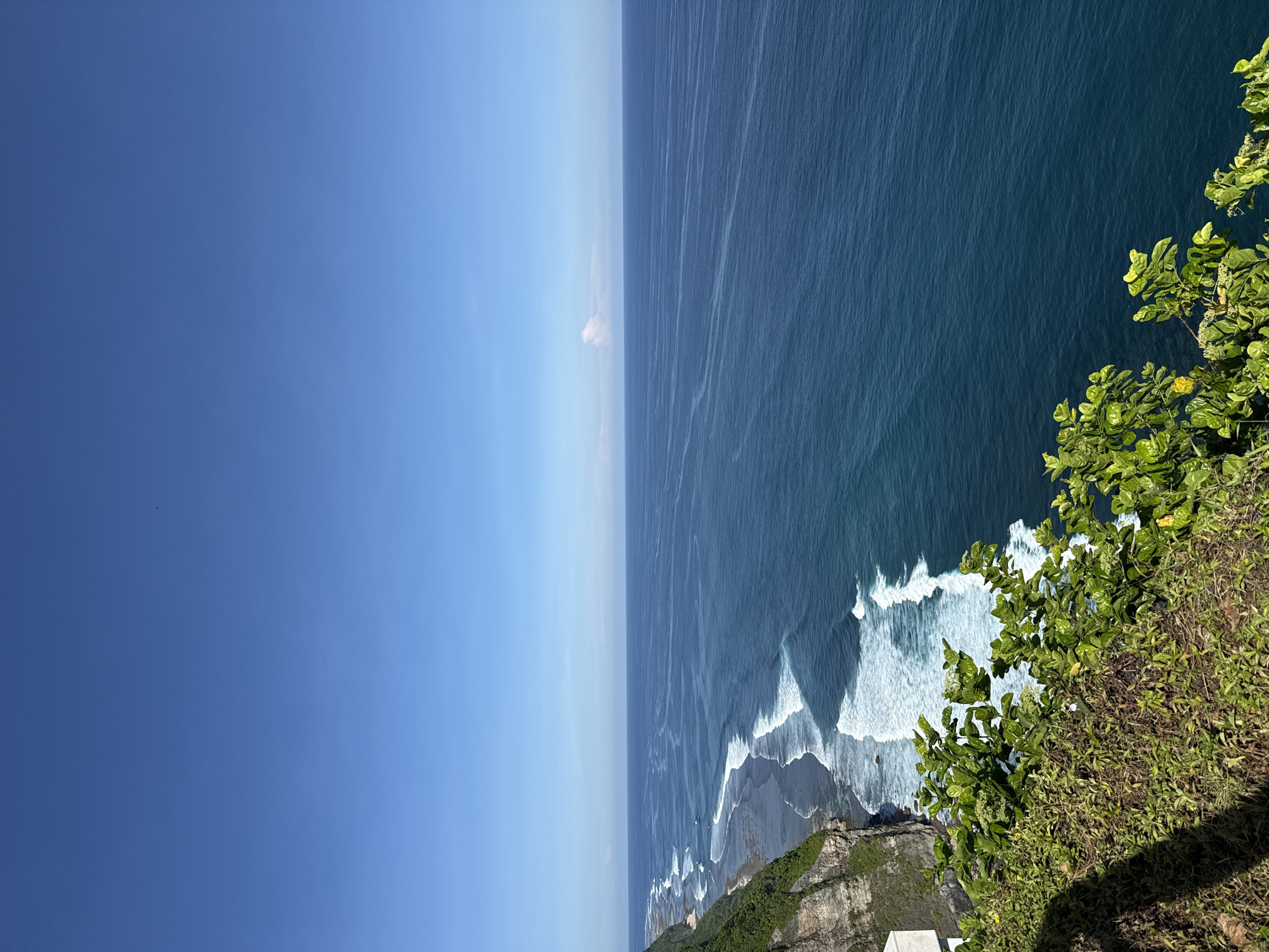 The image shows an aerial view of the coastline in Kuta Selatan, Indonesia. The scene is divided into thirds: a clear blue sky, the deep blue ocean, and a rugged cliffside. Waves break along the base of the cliff, creating white foam against the dark sand. Lush green vegetation covers the cliff's edge. A small structure is visible near the bottom of the cliff.