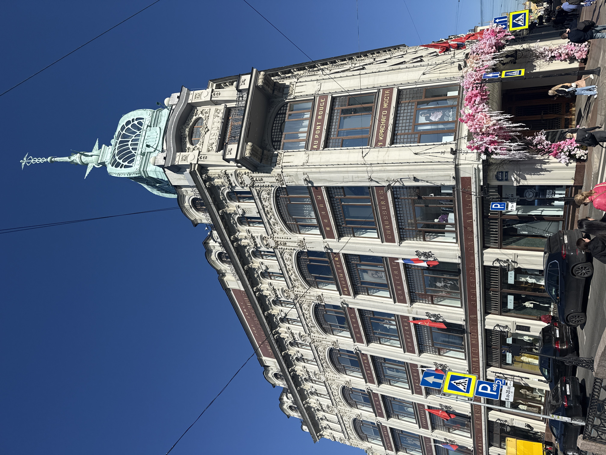 This image displays a grand, ornate building in Saint Petersburg, Russia, under a clear, bright blue sky. The multi-story structure features a light-colored, richly decorated facade with intricate moldings and a mix of arched and rectangular windows, all framed in dark wood.
A prominent feature is the building's rooftop, crowned with a distinctive light-green dome-like structure with a glass roof, topped by a tall, slender spire bearing a caduceus symbol.
Various signs are visible on the building's windows and facade. English and French text includes "AU PONT ROUGE," "LINGERIE," "CHAPEAUN," "CANTERIE," and "CHAUSSURES." Cyrillic text reads "У КРАСНАГО МОСТА" (meaning "At the Red Bridge"), "ЭСДЕРС Ъ СЫНЪ," "КЕФАЛЬСЬ," and "МОЙКИ." The years "1906" and "1907" are also inscribed on the facade. Small Russian flags (red, white, and blue) are displayed from several windows.
The main entrance is elaborately decorated with abundant pink and white artificial floral garlands, forming an archway. At street level, several dark-colored cars are parked along the curb. A few pedestrians are visible on the sidewalk, some near the building's entrance. Street signs include blue pedestrian crossing signs and a "P" parking sign indicating hours from "8.00-20.00." Overhead wires stretch across the clear sky. The scene depicts a bright, sunny daytime.