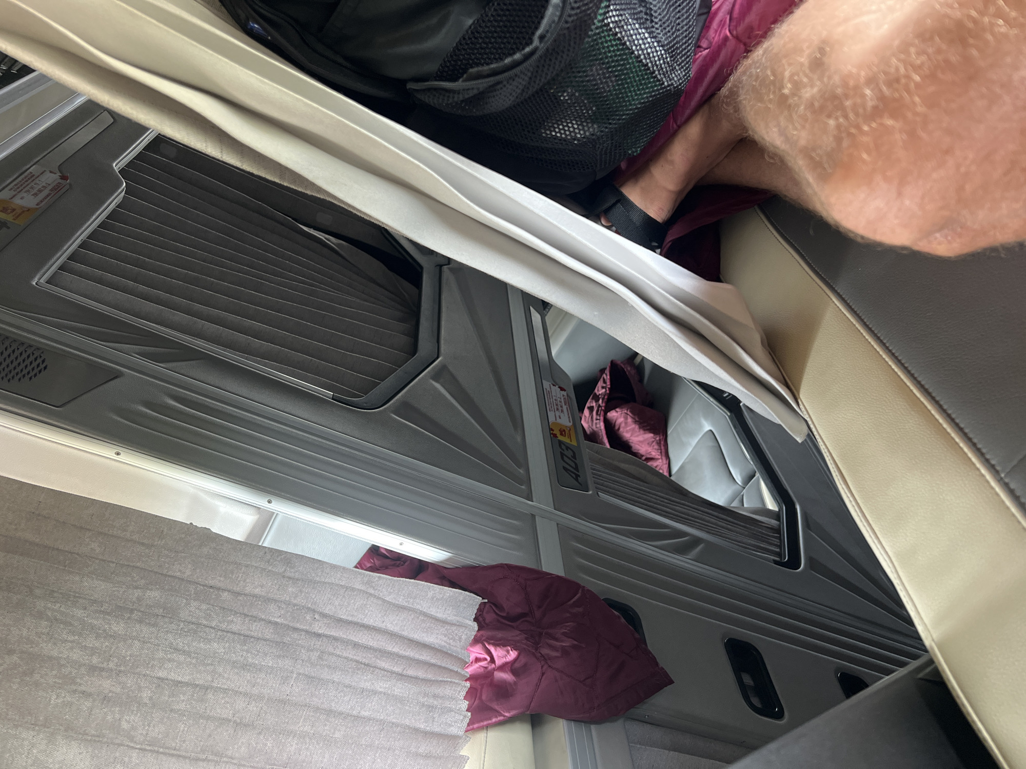 The scene presents an interior view of what appears to be a compartment within a bus or recreational vehicle. Predominantly gray interior panels with horizontal ridges line the walls. A beige and gray seat is partially visible on the right side. A leg, covered in light-colored hair and wearing a black sandal, rests partially on the seat. A black backpack or bag is situated above the compartment, with a burgundy jacket or blanket nearby. A lighter-colored curtain is partially drawn, obscuring the view further into the space. The image does not provide any indication that it is located in Kon Ra Kiah, Vietnam.