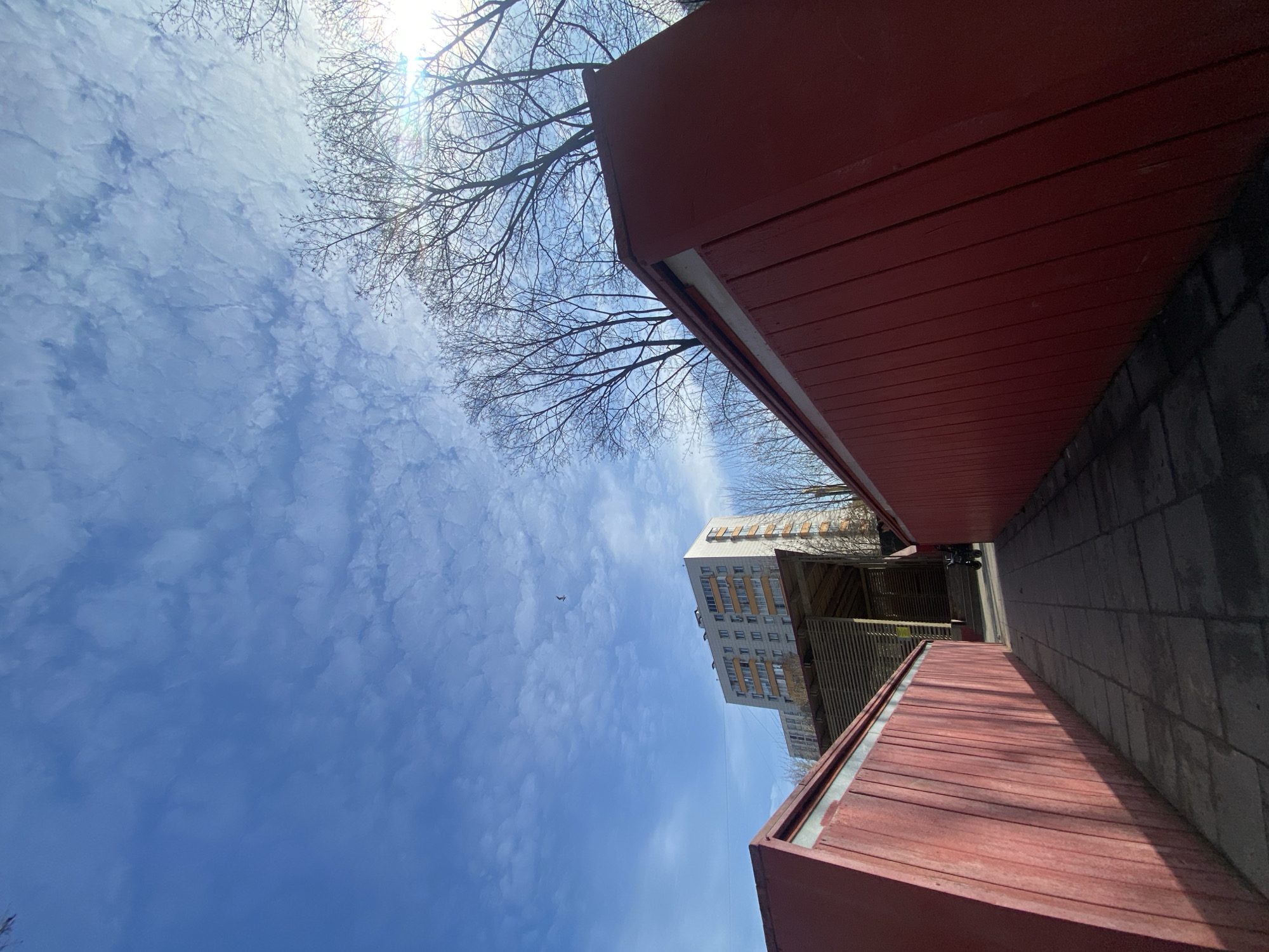 The image shows a view looking up at a partially cloudy blue sky, framed by the edges of red buildings or structures on either side. Bare tree branches are visible against the sky. In the distance, a multi-story building is visible, as well as some kind of walkway or structure with a slatted design. The image was taken in Khimki, Russia.