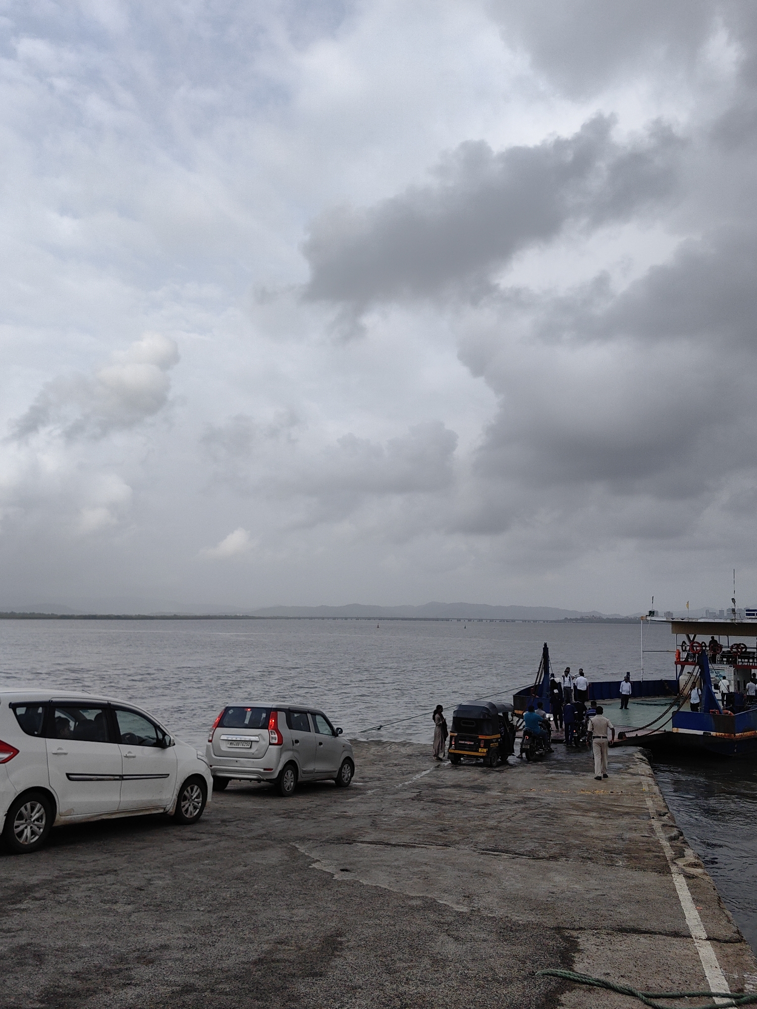 The image captures a scene at a ferry terminal in Mira-Bhayander, India, on an overcast day. In the foreground, a paved concrete surface slopes gently towards the water. Two cars are parked on this surface: a white multi-purpose vehicle on the left and a silver hatchback on the right, facing away from the viewer.
Further back, at the edge of the concrete, a ferry is docked. Several people are visible on the ferry's deck and on the ramp connecting it to the shore. Among them are individuals in white shirts, suggesting they might be crew members. A three-wheeled auto-rickshaw is also present near the ferry.
The environment is characterized by a large body of water stretching towards the horizon, where a distant landmass with hills is visible. A faint bridge or structure can be seen in the far distance across the water. The sky is filled with heavy, grey clouds, indicating a cloudy or possibly rainy day. The overall atmosphere is subdued due to the weather conditions.