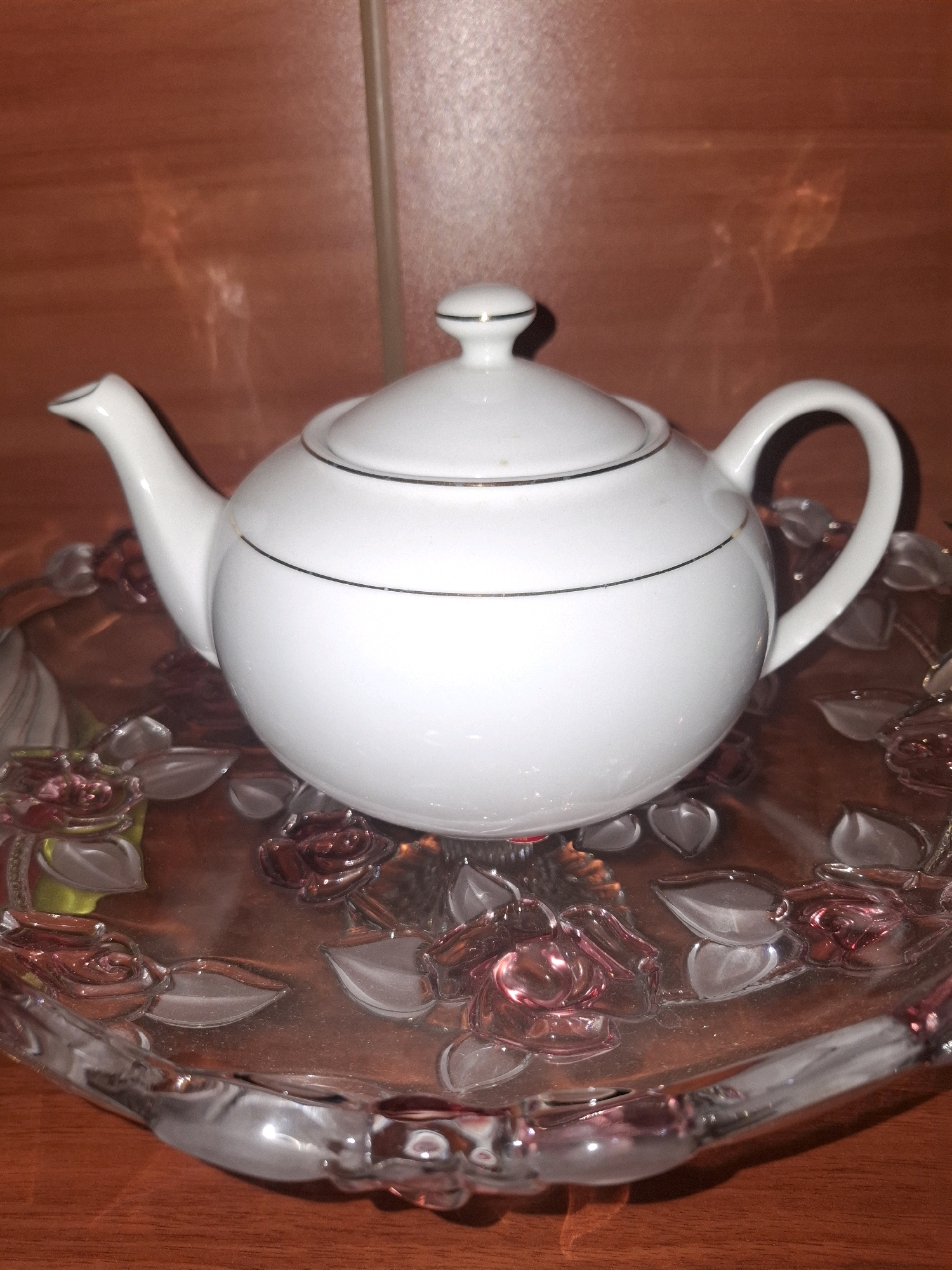 A white ceramic teapot with gold trim sits on a decorative glass plate. The plate is clear with raised floral designs in shades of pink and red. The teapot has a lid with a knob, a spout, and a handle. The background is a wooden surface with a textured pattern. The scene appears to be indoors, possibly on a table. The image does not contain any recognizable landmarks or elements that would indicate its location in Almaty, Kazakhstan.