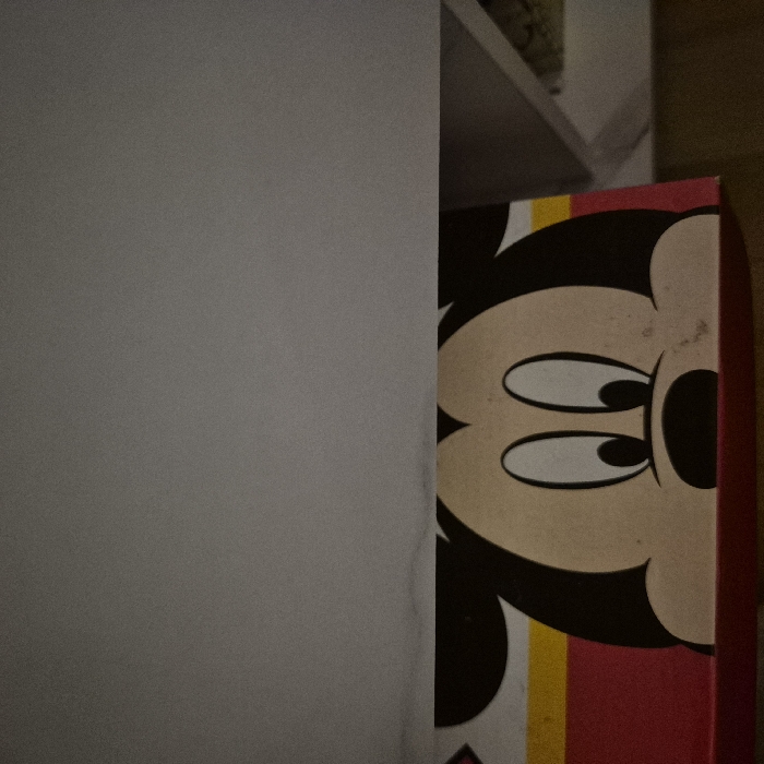 A partial view reveals a depiction of Mickey Mouse. The character’s face is tan, with black ears, white eyes, and a black nose. The image is vertically oriented, with the top of Mickey's head near the bottom of the frame. Red, yellow, and white striped lines are present behind the character. The image appears to be on a box or container. Part of a shelf is visible. The scene is illuminated by dim lighting.