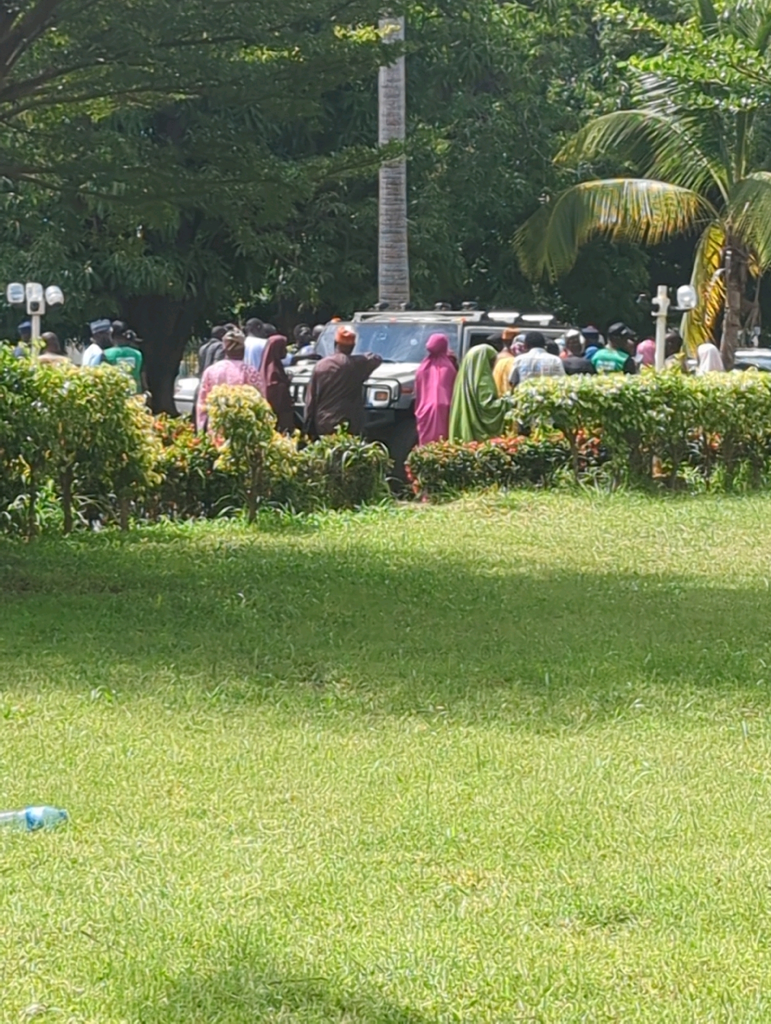 In Unguwan Dosa, Nigeria, a group of people are gathered near a white vehicle on a street lined with dense green foliage and trees. Several individuals are visible wearing traditional attire, including a man in a brown garment and a red cap, and women in brightly colored hijabs, one pink and one green. The foreground of the image features a well-maintained lawn with a clear plastic bottle lying on the grass. The background is dominated by lush green trees and palm fronds, suggesting a tropical or subtropical environment. The scene appears to be a public gathering or event.