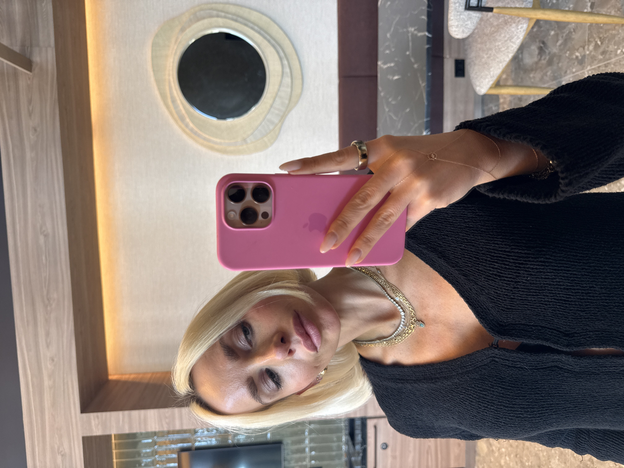 A woman is taking a selfie with a pink iPhone. She has blonde hair, is wearing a black top, and several necklaces. She also has a gold ring and a hand chain. The background includes a wall with a round decorative mirror, a chair, and wooden paneling.