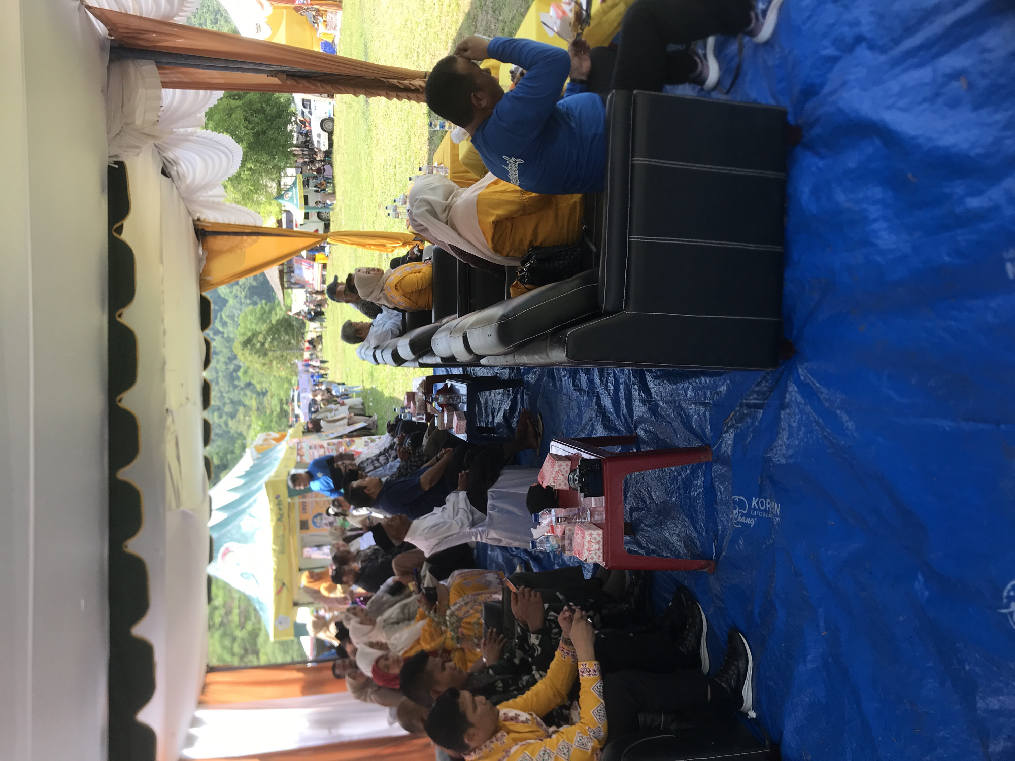 The image depicts an outdoor event in Langkuta, Indonesia, under a large tent. In the foreground, a group of people are seated on black couches and chairs, some on a blue tarp. One man in a yellow shirt is looking at his phone, while another in a blue shirt is touching his head. To the right, a man in a blue long-sleeved shirt with white writing is visible, as well as a yellow table with bottles of water and boxes. In the background, more attendees are seated, and tents with yellow and blue accents are set up on a grassy field. Beyond the field, trees and hills are visible. The overall scene suggests a community gathering or event.