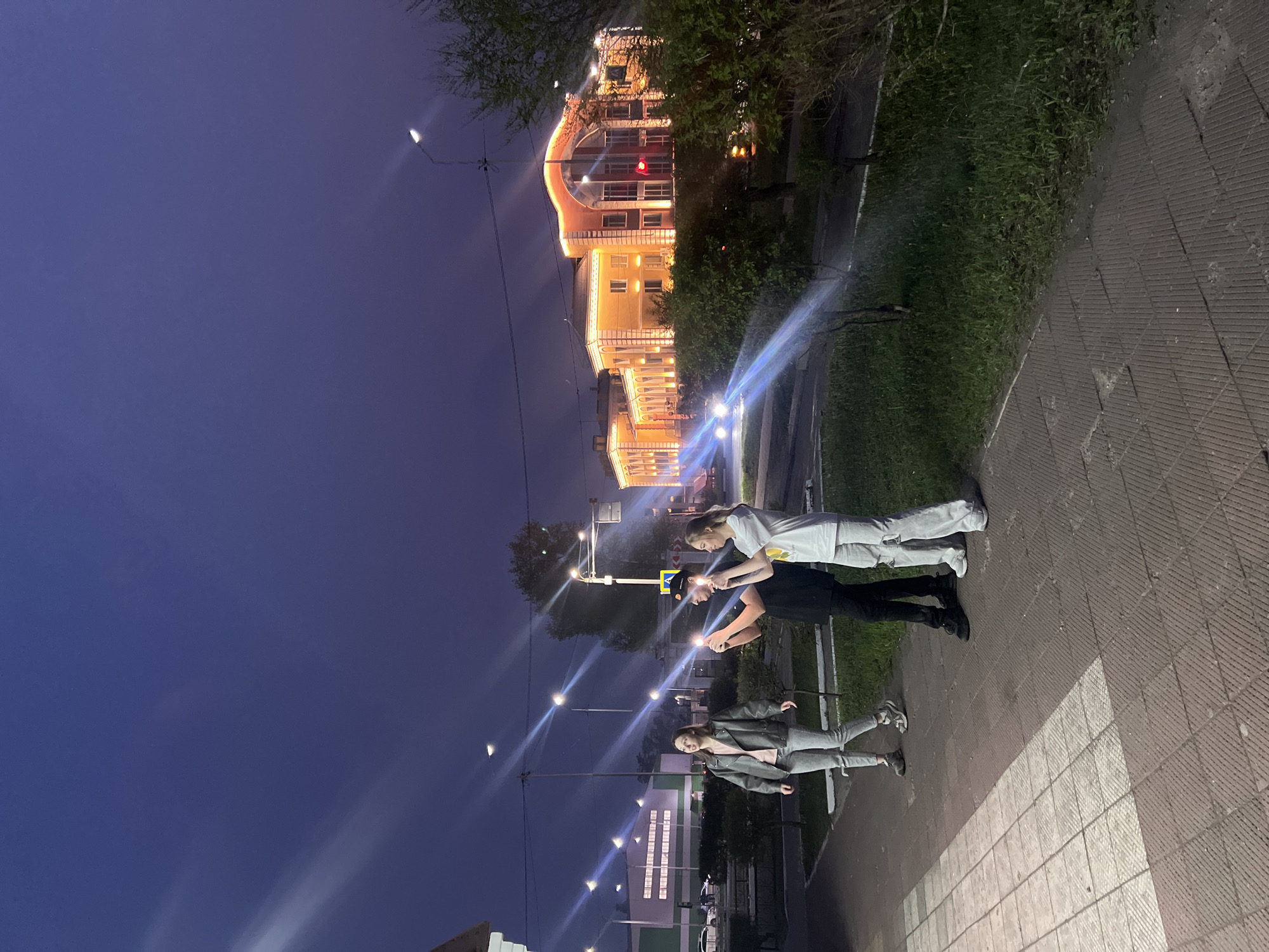 The image shows an urban scene at night. In the center, a man in a black shirt and baseball cap is holding a phone, and a woman in a white top appears to be assisting with the phone or looking at its screen. Another woman, wearing a grey jacket and pants, is standing to the left. In the background, a large, lit building is visible, along with streetlights and greenery. The group of people are standing on a tiled sidewalk.