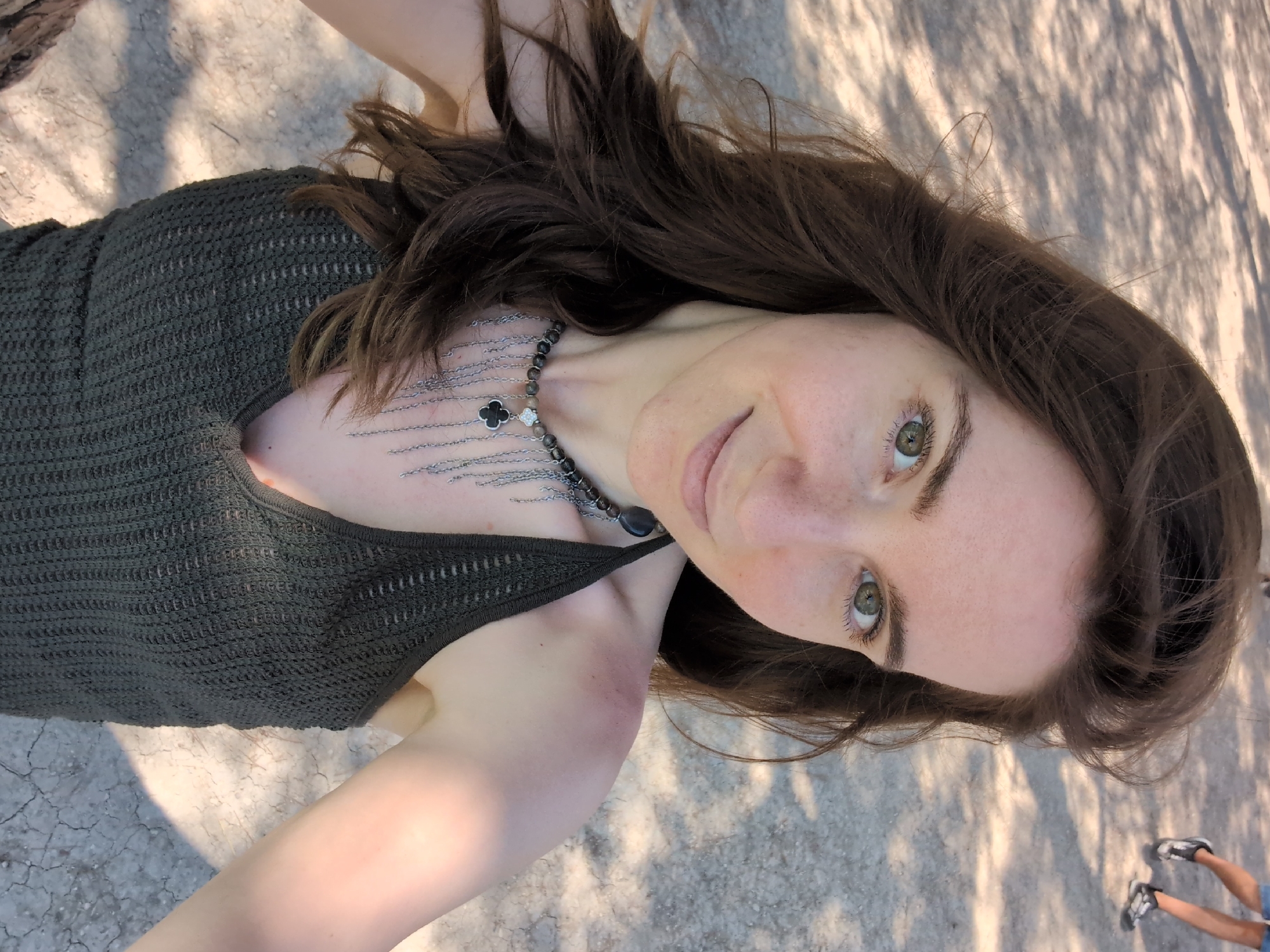 A young woman with long, brown hair and green eyes is taking a selfie outdoors. She is wearing a dark green, knitted tank top and a necklace with dark beads and a clover pendant. Her skin is fair with freckles. The background is a sandy or gravelly ground with dappled sunlight and shadows. In the upper right corner of the image, the legs of another person, wearing blue shorts and athletic shoes, are visible. The location is indicated to be the city of Divnomorskoye, Russia.