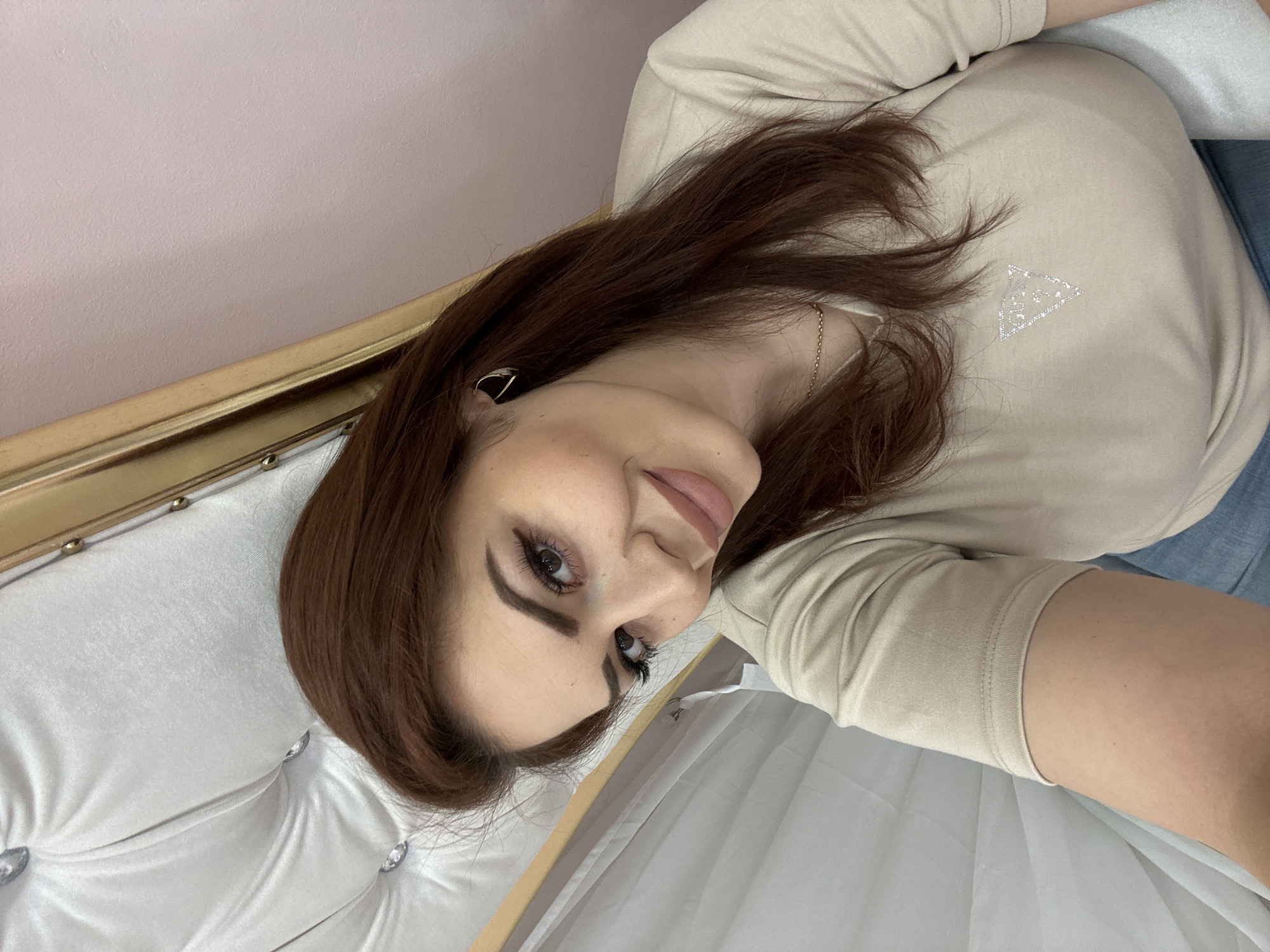 A woman with brown hair is lying down on a white tufted headboard with gold trim. She is wearing a light beige long-sleeved shirt with a triangle logo on the chest, and her skin is light-toned. She is wearing a gold earring and a delicate gold necklace. The wall behind the headboard is a light pink color. The setting appears to be indoors. There is no information available to suggest that this image was taken in the city of Sofia, Bulgaria.