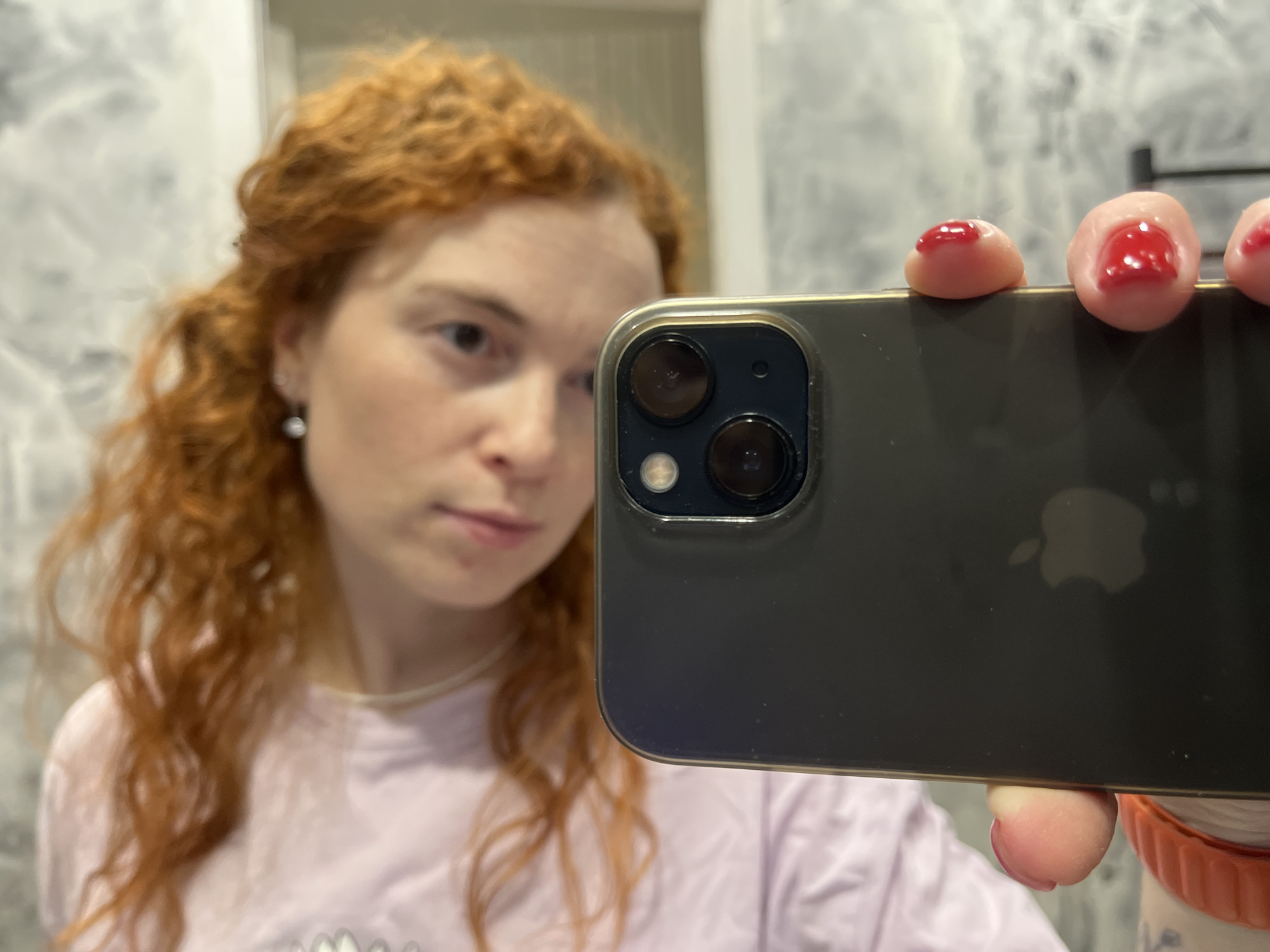 A close-up selfie captures a woman with prominent red, curly hair and light eyes, taken in Chaikovsky, Russia. She holds a dark-colored smartphone, appearing to be an iPhone given the visible Apple logo on the back and the distinct dual camera system encased in a clear cover, partially obscuring her face as she looks towards the camera. Her fingers, with short, rounded nails painted in bright red polish, are visible gripping the device. She wears a light-colored, possibly pink or lavender, t-shirt and a thin, light-toned necklace. A subtle pearl-drop earring is visible on her left ear. The background is softly blurred, displaying muted shades of white and gray, characteristic of an indoor setting like a bathroom or bedroom. The lighting is bright and even, suggesting indoor artificial light or diffuse daylight. Her expression is neutral and contemplative.
