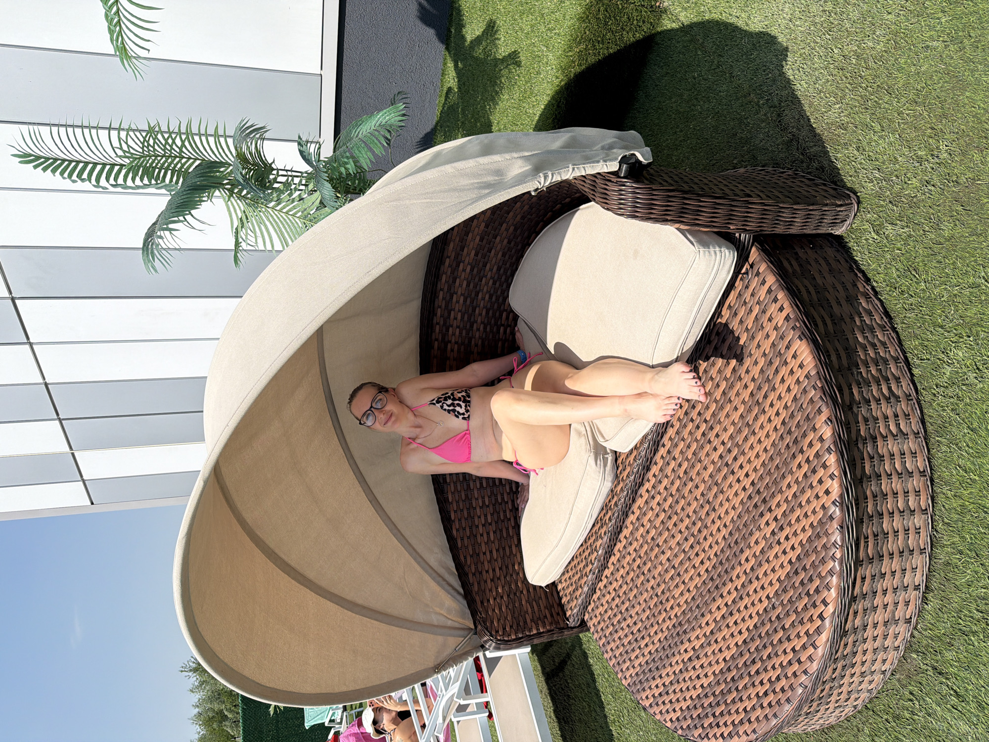 A woman with blonde hair and glasses is reclining on a round, brown wicker chaise lounge with a beige canopy. She is wearing a pink and animal print bikini. The chaise lounge is on green grass, and there is a palm tree visible next to a building. The building is white with gray accents. The sky is blue. The location is likely Moscow, Russia.