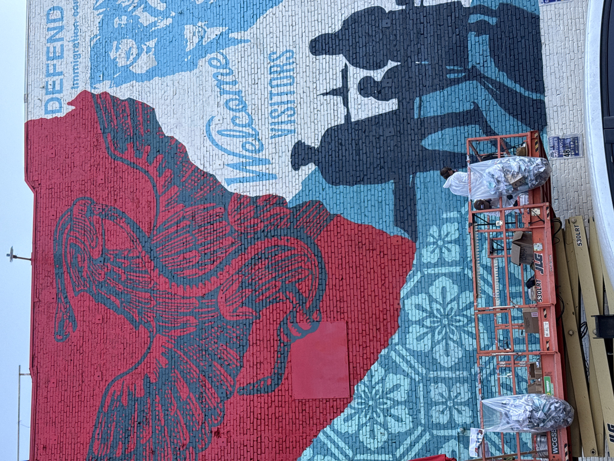 The image shows a large mural painted on a brick wall in an urban setting. The mural features bold colors and imagery, including a red bird-like figure with dark blue accents on the left, and a silhouette of people with the words "WELCOME VISITORS" and "DEFEND Immigration" on the right. Below the mural, an orange scissor lift is positioned, with two people in white protective suits working on it, seemingly engaged in painting or maintenance. Various supplies and boxes are visible on the platform of the lift. The wall appears to be part of a building, and a street or car is visible in the foreground. The sky above is a clear blue. The mural's style suggests a message of welcome and support for immigrants, contrasted with a call to defend immigration. The presence of the scissor lift and workers indicates that the mural is either new or undergoing restoration. The overall scene captures a moment of artistic creation or preservation in an urban landscape.