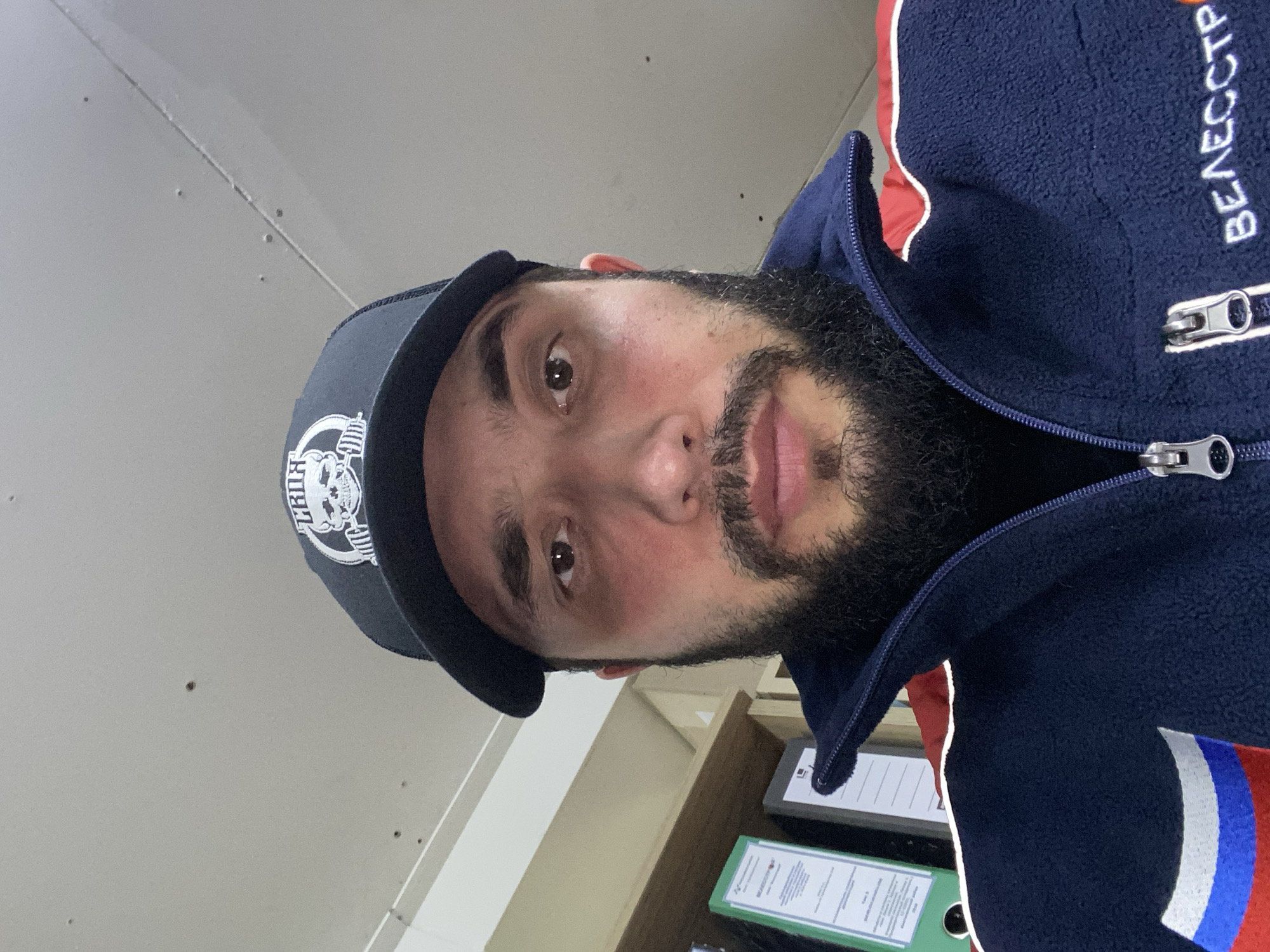 A medium shot captures a man from the chest up, looking directly at the camera. He is wearing a black baseball cap with a white logo featuring a skull and barbells. The man has a dark beard and a dark complexion. His jacket is navy blue with red and white accents on the collar and a white text on the right side of the chest. Behind him, to his left, is a shelf containing several folders, one of which is bright green and has visible text. The background appears to be an indoor setting with a light-colored ceiling and wall. The overall impression is a casual portrait, possibly taken in an office or personal workspace in Luzhitsy, Russia.