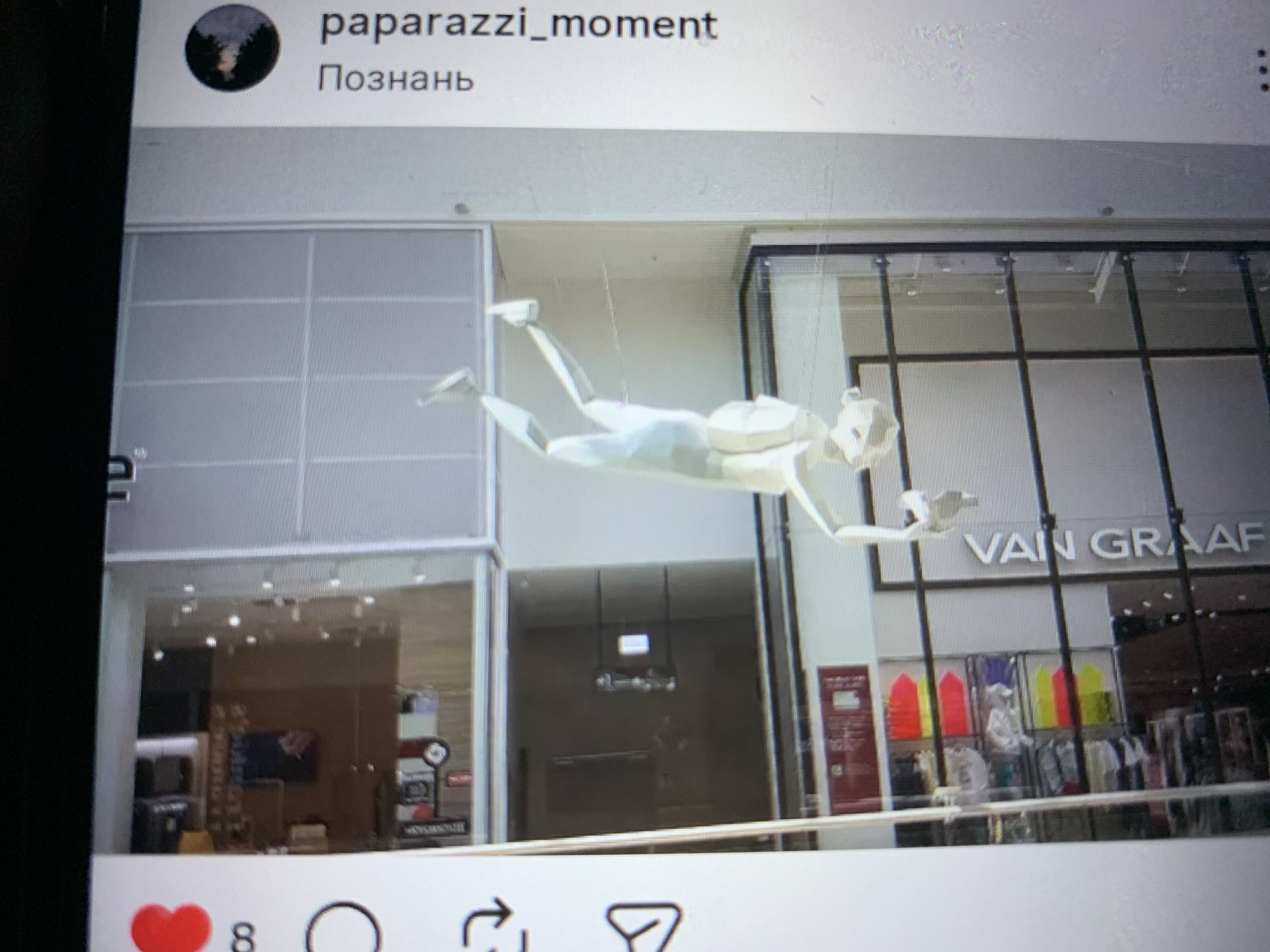 The image shows a store interior with a mannequin hanging upside down. The mannequin is white and appears to be made of plastic or fiberglass. It is positioned in the center of the frame, with its arms extended and its legs pointing upwards. The mannequin is wearing a white outfit. In the background, there are shelves stocked with various items, including colorful clothing. The store appears to be a retail establishment, possibly a clothing store, given the merchandise visible. There are some text elements in the image, but they are not clearly legible. The lighting in the store is bright, illuminating the mannequin and the merchandise. There is no indication of the location in the city of Lymanka, Ukraine. The image is a photograph taken from a slightly elevated angle, possibly by a customer or an employee.