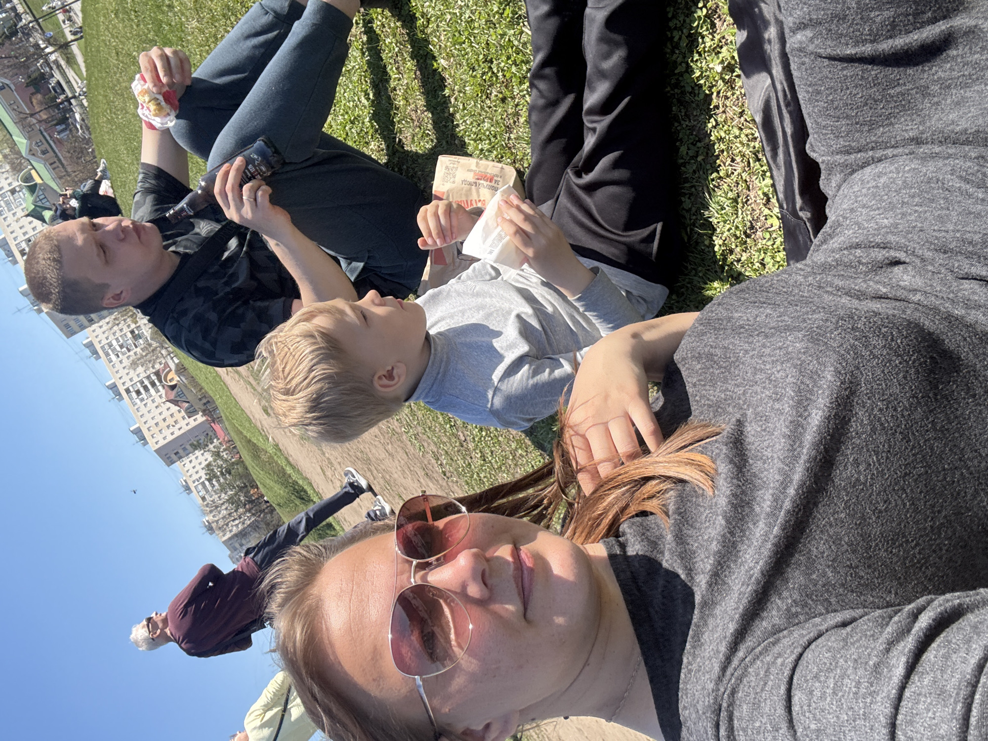 The image shows a group of people lying on the grass in Dmitrov, Russia. A woman wearing sunglasses is closest to the camera, reclining with her head resting on the ground. A young boy lies beside her, looking at a fast-food item in his hands. A man lies next to the boy, holding a beer bottle and a snack. Buildings are visible in the background. An older man walks by in the background. The weather appears to be sunny.
