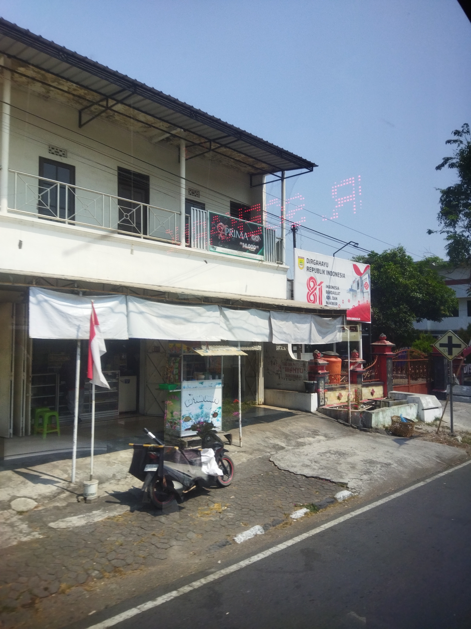 In the city of Sukoharjo, Indonesia, a white building with two stories stands prominently. The upper floor features a balcony with a silver railing and two windows with dark frames. Below, the ground floor is a commercial space with roll-up doors, partially open, revealing storefronts. A white banner hangs across the front of the ground floor, and a red and white Indonesian flag is displayed on a pole.
On the right side of the building, a large banner with text that includes "DIRGAHAYU REPUBLIK INDONESIA" and "81" is visible. Above the building, large, bright pink digital letters, possibly part of a sign, are displayed against the clear blue sky.
In front of the building, a sidewalk or paved area is present. A dark-colored motorcycle with a cargo box is parked on the pavement. A small food cart with a colorful graphic depicting fish is also present. Further to the right, a decorative concrete structure with planters and an orange water tank is situated. A red metal fence encloses an area behind this structure. A traffic sign with a yellow cross symbol is visible on a pole to the far right. The scene suggests a street-level view in a commercial or residential area.