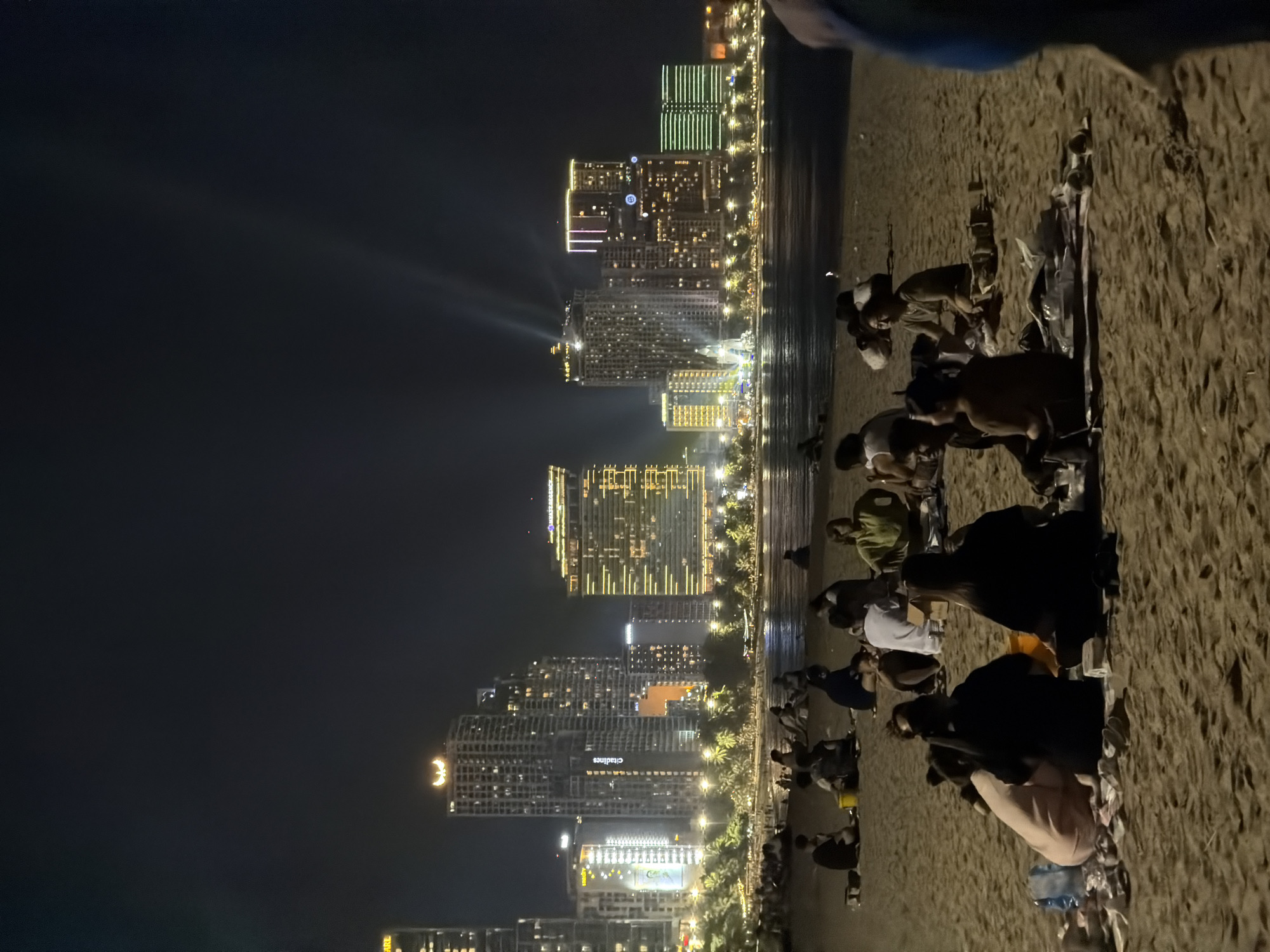 The image depicts a nighttime scene at Nha Trang Ward, Vietnam. In the foreground, multiple groups of people are seated on a sandy beach, appearing to be engaged in eating or socializing. The background features a cityscape with numerous high-rise buildings illuminated with various lights, creating a vibrant urban backdrop against the dark sky. The beach stretches alongside the city, forming a visible boundary between the urban environment and the natural coastline. The light reflects off the water onto the sand.